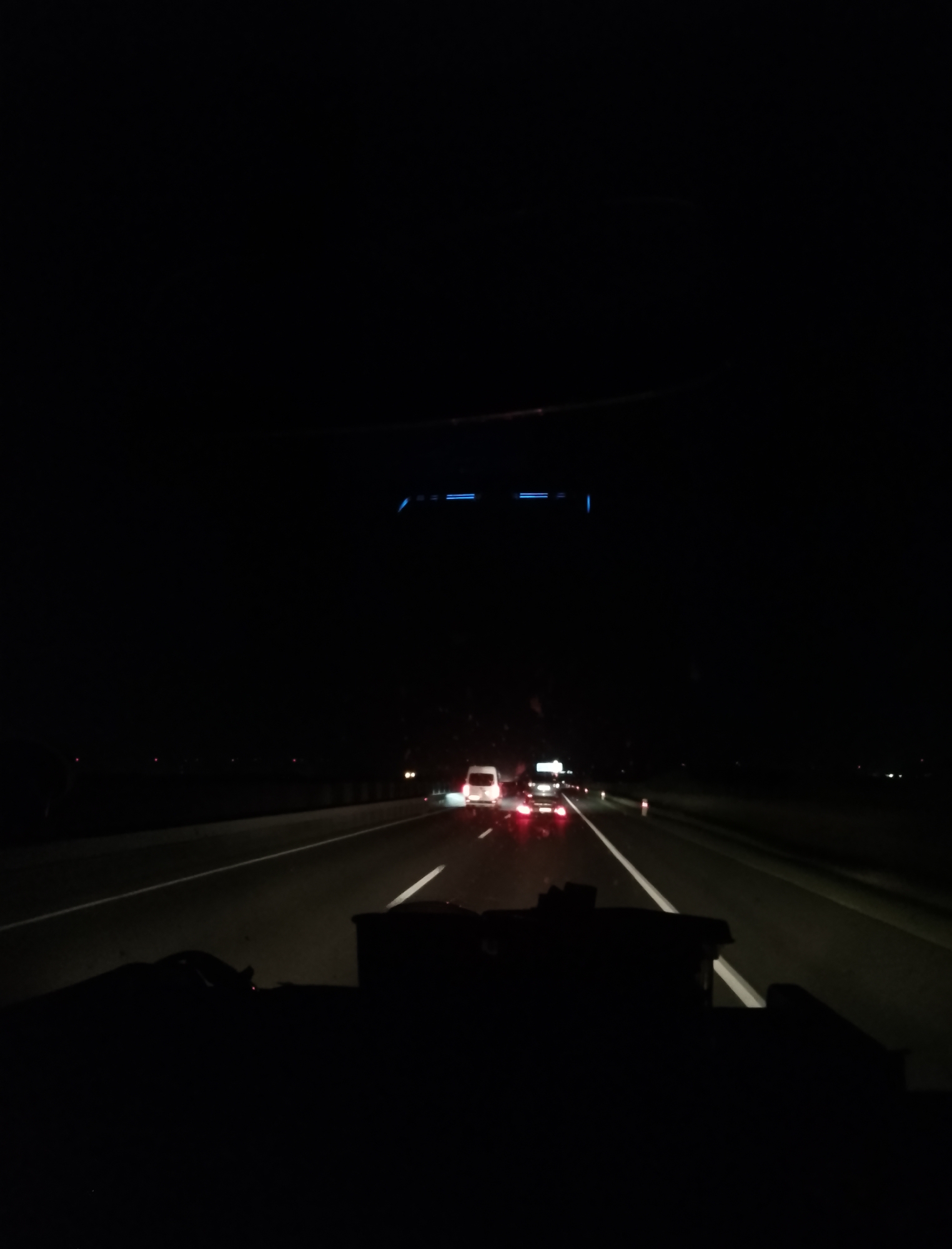 The image depicts a nighttime view from inside a vehicle traveling on a highway. The perspective is from the driver's seat, looking forward. The road ahead is illuminated by the headlights of other vehicles and streetlights, creating a stark contrast with the surrounding darkness. Several cars are visible on the highway, their taillights and headlights glowing. The road itself has white lane markings. In the foreground, the dashboard and parts of the vehicle's interior are silhouetted against the dimly lit road. The sky above is black, indicating it is late at night or very early morning. There is no identifiable cityscape or specific location visible, suggesting an open highway environment rather than a dense urban area. The blue light reflected on the windshield suggests the presence of some electronic display or ambient light source within the vehicle's cabin.