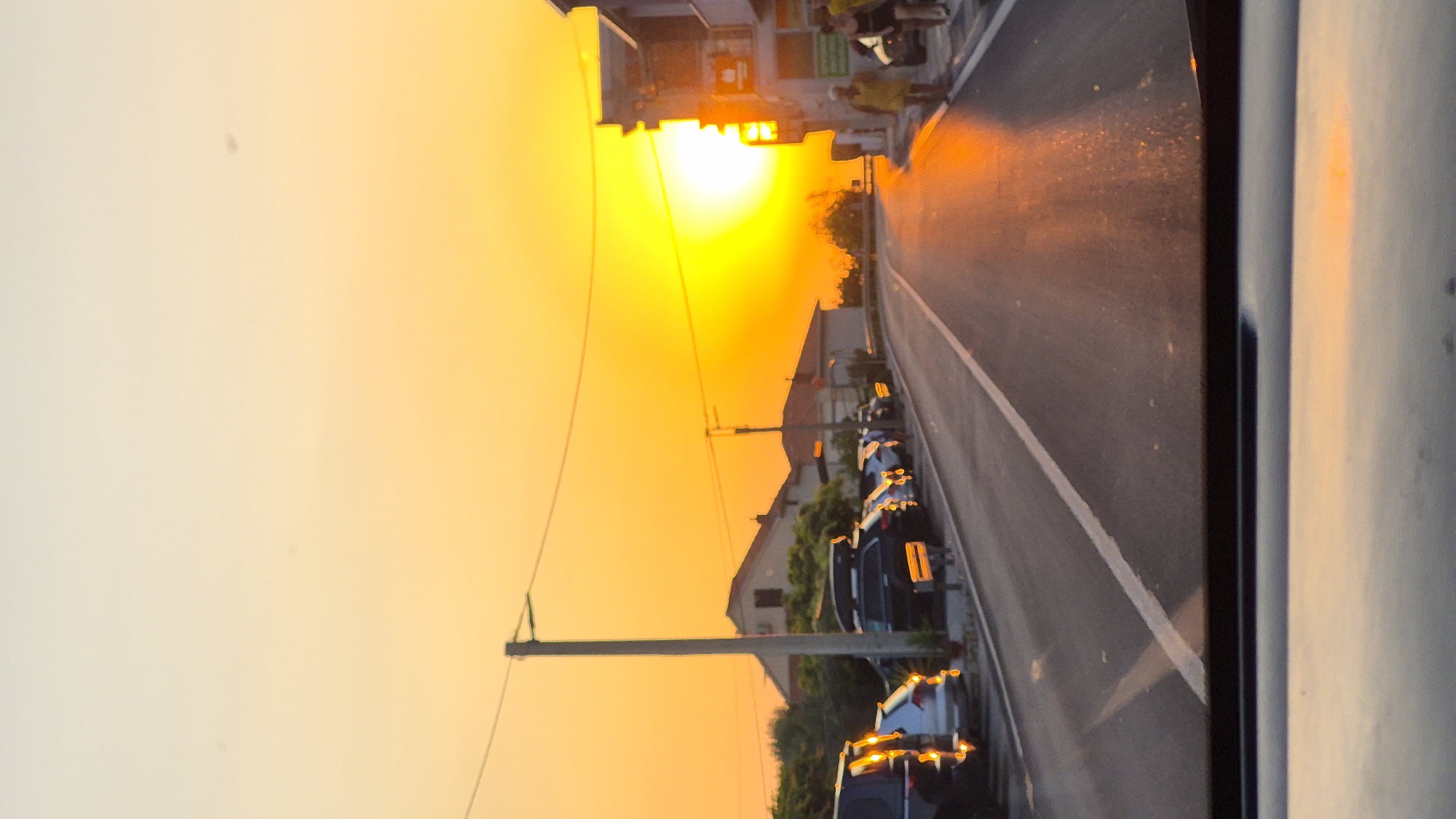 A vertical, full-shot, eye-level, wide-angle exterior photograph captures a street scene at sunset in an unknown location. The sky is a brilliant, saturated orange-yellow, with the sun prominently visible on the right side of the frame, appearing as an intensely bright, slightly blurred orb. The sun's glow illuminates the entire scene with warm tones.
In the foreground, the reflection of the sunset is visible on what appears to be a car's dashboard or windshield. The road stretches into the distance, paved with asphalt and marked with a white dividing line. The road surface is wet, with patches of light reflecting the intense sunset.
To the left of the road, a row of parked cars is visible. The cars are of various types and colors, some with their headlights or taillights illuminated. Further back, residential buildings with red-tiled roofs and green foliage are present. A utility pole stands near the left side of the road, with power lines stretching across the frame.
On the right side of the road, a building with multiple stories is visible, featuring windows and balconies. Two individuals are walking on the sidewalk in front of this building, silhouetted against the bright sky. The overall impression is that of a serene and picturesque sunset over a street in a residential or small town area.
