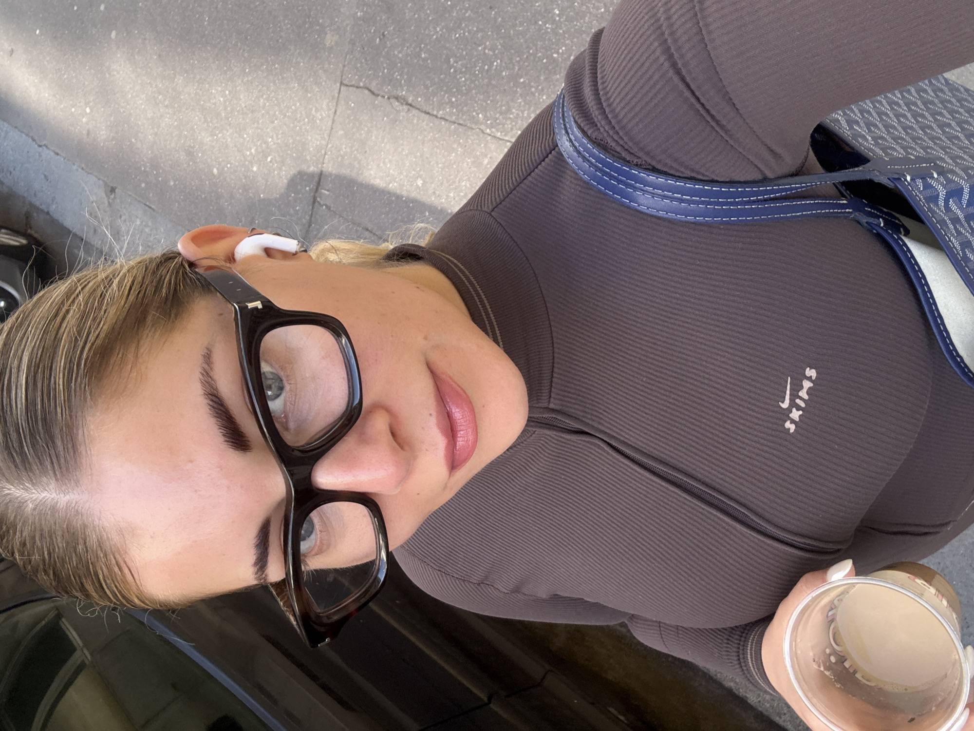A woman with blonde hair and glasses holds a coffee cup in her right hand, her fingernails painted white. She wears a fitted, dark brown, ribbed long-sleeved shirt with a zipper that is partially visible. The word "SKIMS" is visible in light tan on the left chest of the shirt. In her left ear, she wears a white earbud. The strap of a blue and white patterned bag is over her shoulder. She is standing on a city street. The location is Paris, France.