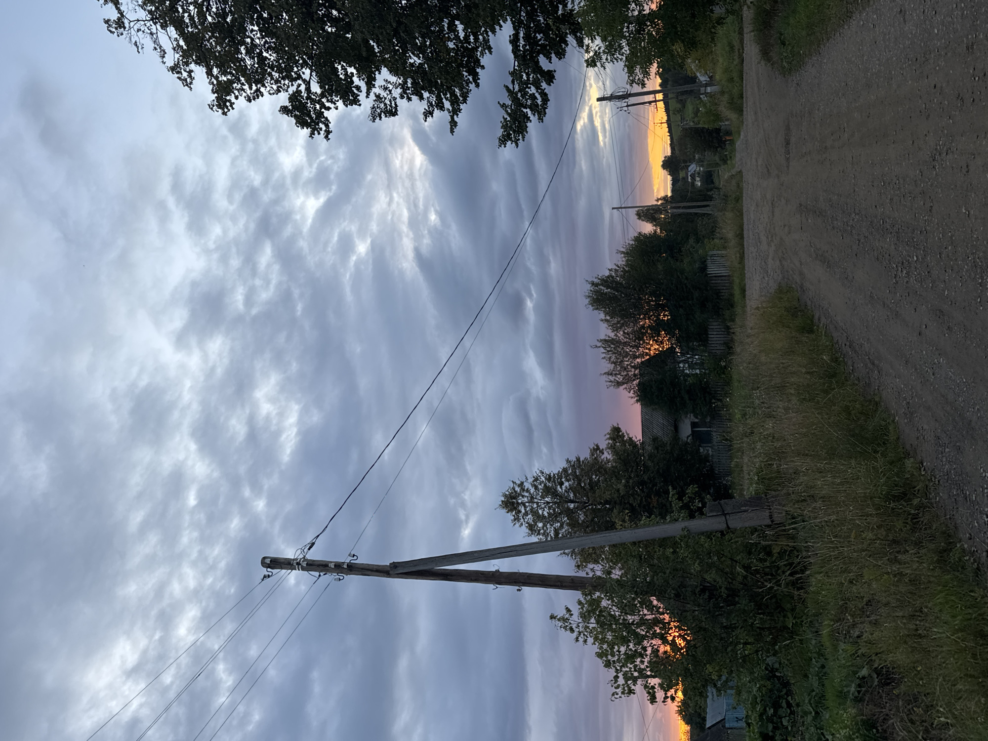 The image depicts a rural scene in the village of Borodulino, Russia, at dusk. A dirt road, appearing to be an intersection, dominates the foreground and leads into the distance. To the left of the road, tall grass and trees are visible, with a wooden power pole standing prominently. In the background, houses and more trees are scattered across the landscape. The sky is filled with dramatic clouds, with a warm orange and pink glow near the horizon, indicating the setting sun. The overall atmosphere is one of quiet evening in a rural setting.