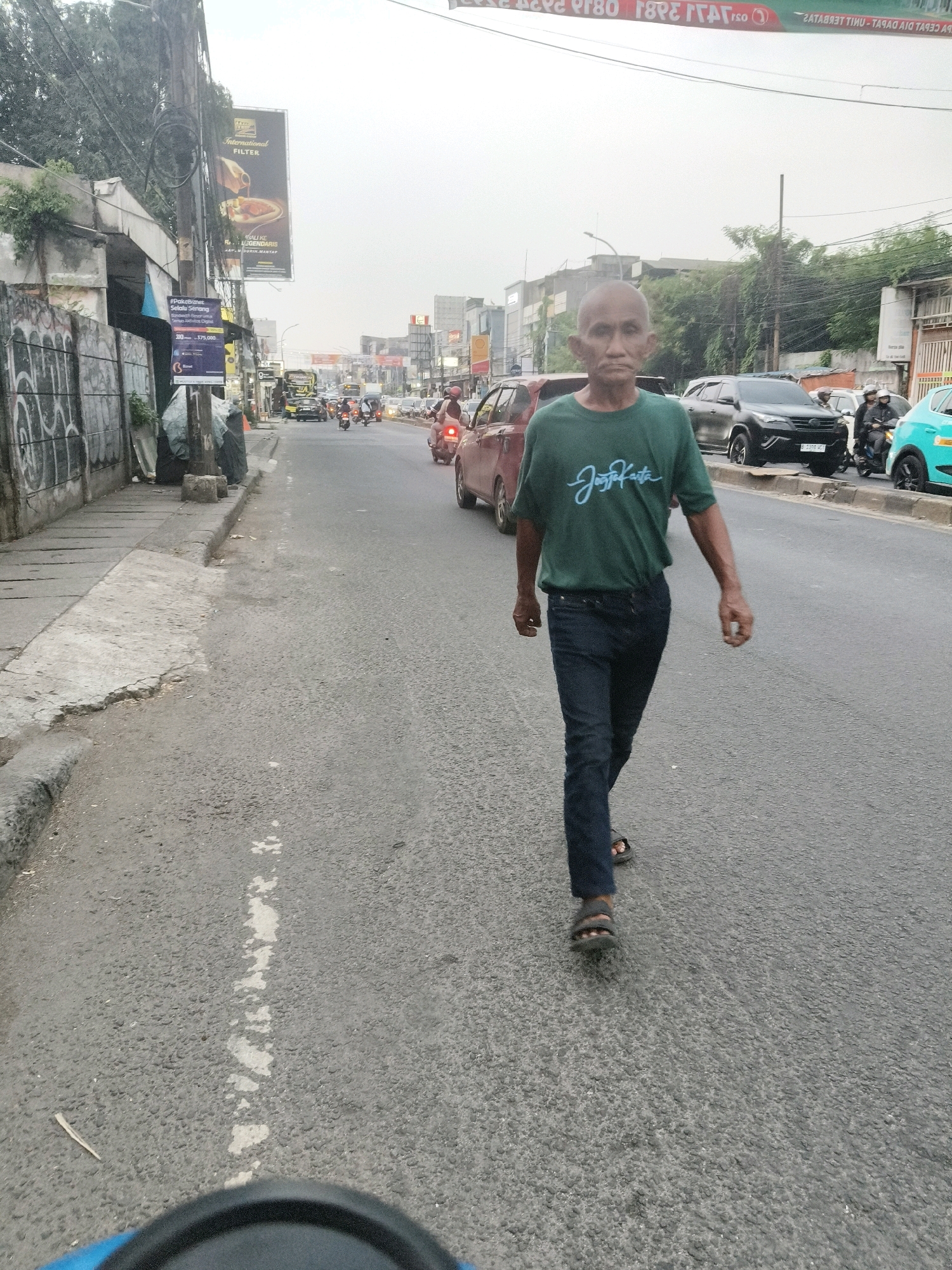 A man walks on a road in South Tangerang, Indonesia, wearing a green t-shirt and dark pants. He is bald and appears to be of middle age or older. He is walking towards the camera on the right side of the road, which has a sidewalk on the left. There are several vehicles on the road, including cars, motorcycles, and a bus. The environment appears to be urban, with buildings and streetlights visible in the background. The sky is overcast.