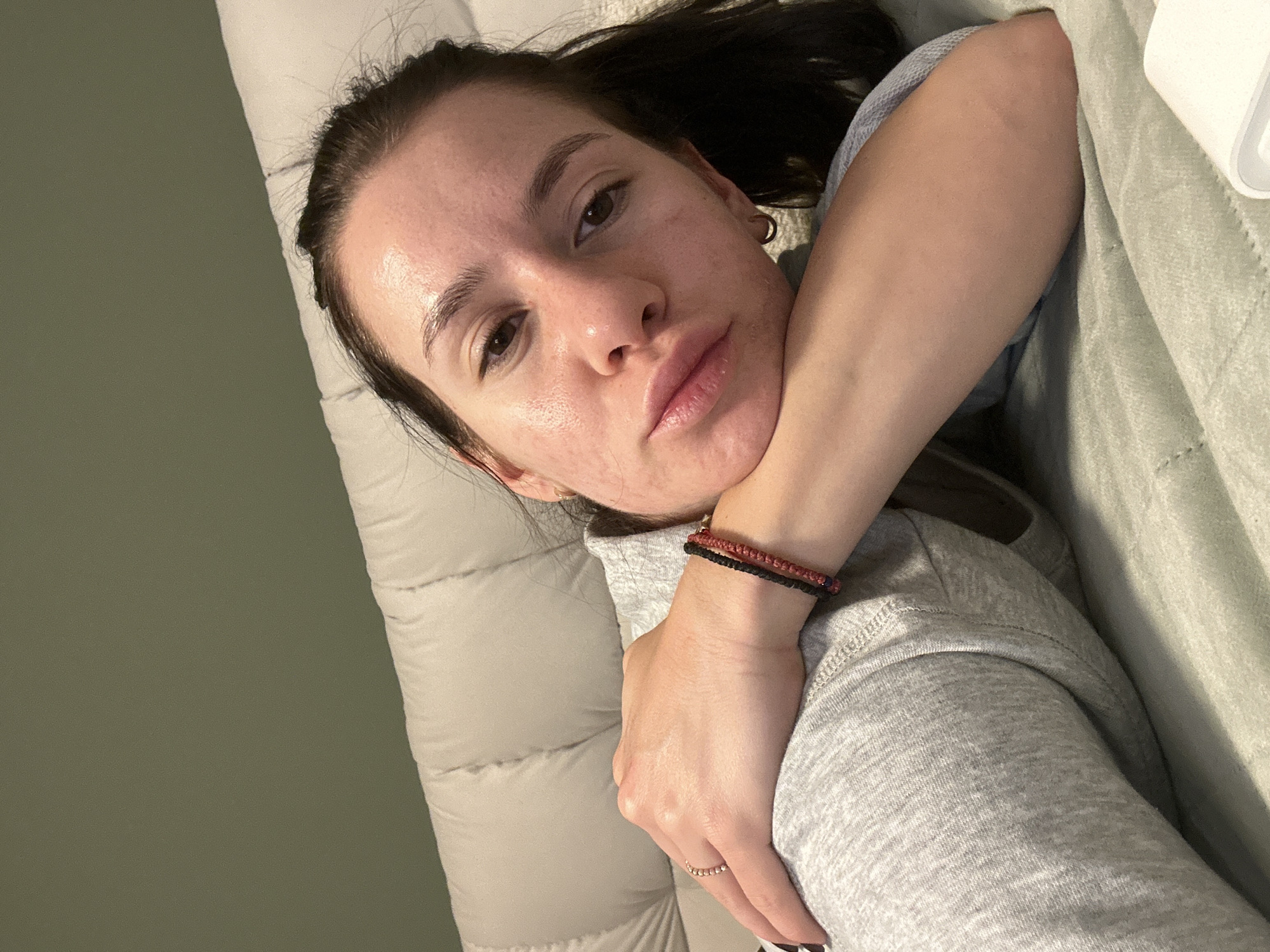 The image is a close-up, eye-level portrait of a young adult woman lying down indoors. She is positioned on her side on a light-colored, quilted surface, likely a bed or sofa, with an olive-green wall visible behind her. Her dark hair is pulled back, revealing her face which has visible skin texture and a slightly glossy forehead. She has brown eyes, full lips, and a neutral, direct gaze towards the camera, suggesting a selfie or self-portrait.
She is wearing a casual light grey long-sleeved top, possibly a hoodie. Accessories include a small gold hoop earring in her left ear, two thin woven bracelets (one red, one black) on her right wrist, and a delicate gold-toned ring with small beads on her right ring finger. Her arms are bent, with her hands resting near her chest and neck. The lighting is soft and ambient, indicative of an indoor setting, possibly in the evening. The scene depicts a relaxed moment indoors in Plovdiv, Bulgaria. No visible text is present.
