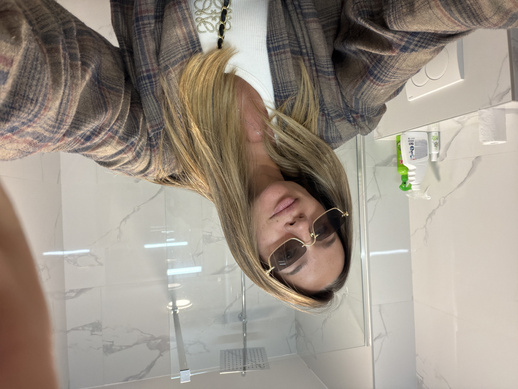 A selfie taken in a bathroom setting with a woman wearing a plaid blazer over a white top. She is also wearing sunglasses. The bathroom has white marble walls and a shower stall with a chrome showerhead. The location is the city of Unknown, Unknown.