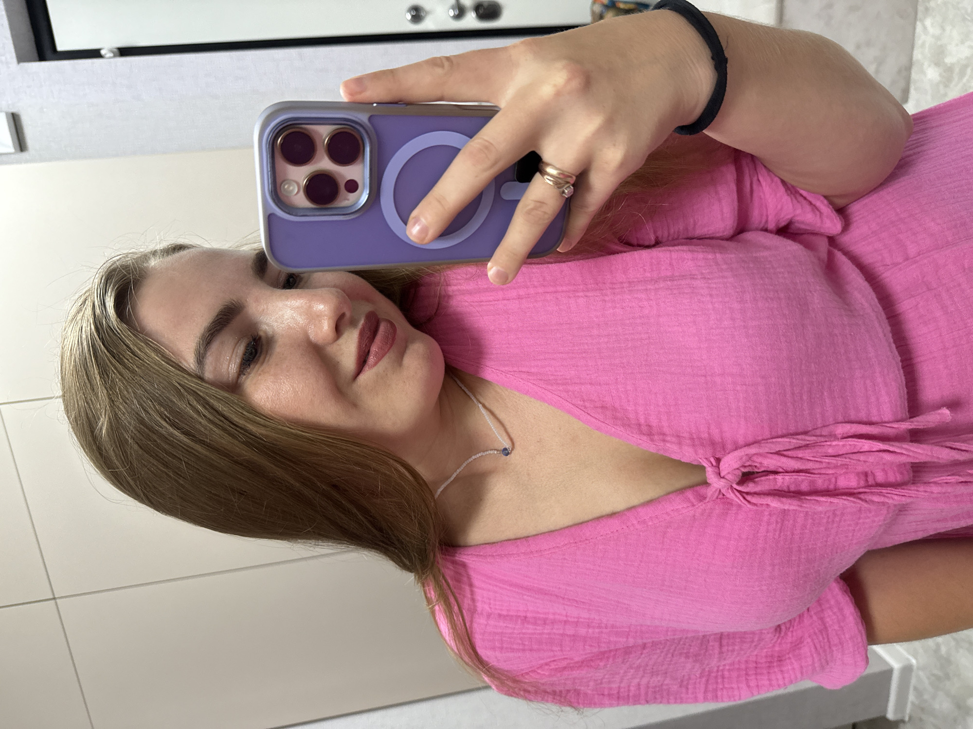 A young woman with long, blonde hair takes a selfie in a room with light-colored walls and a mirror. She is wearing a pink, v-neck dress with a tie front and short sleeves. She has a necklace with a small blue pendant and is wearing two rings on her left hand. She is holding a purple iPhone with a case that has a circular magnetic charging area. The background shows a mirror reflecting part of the room, including a door with a handle.