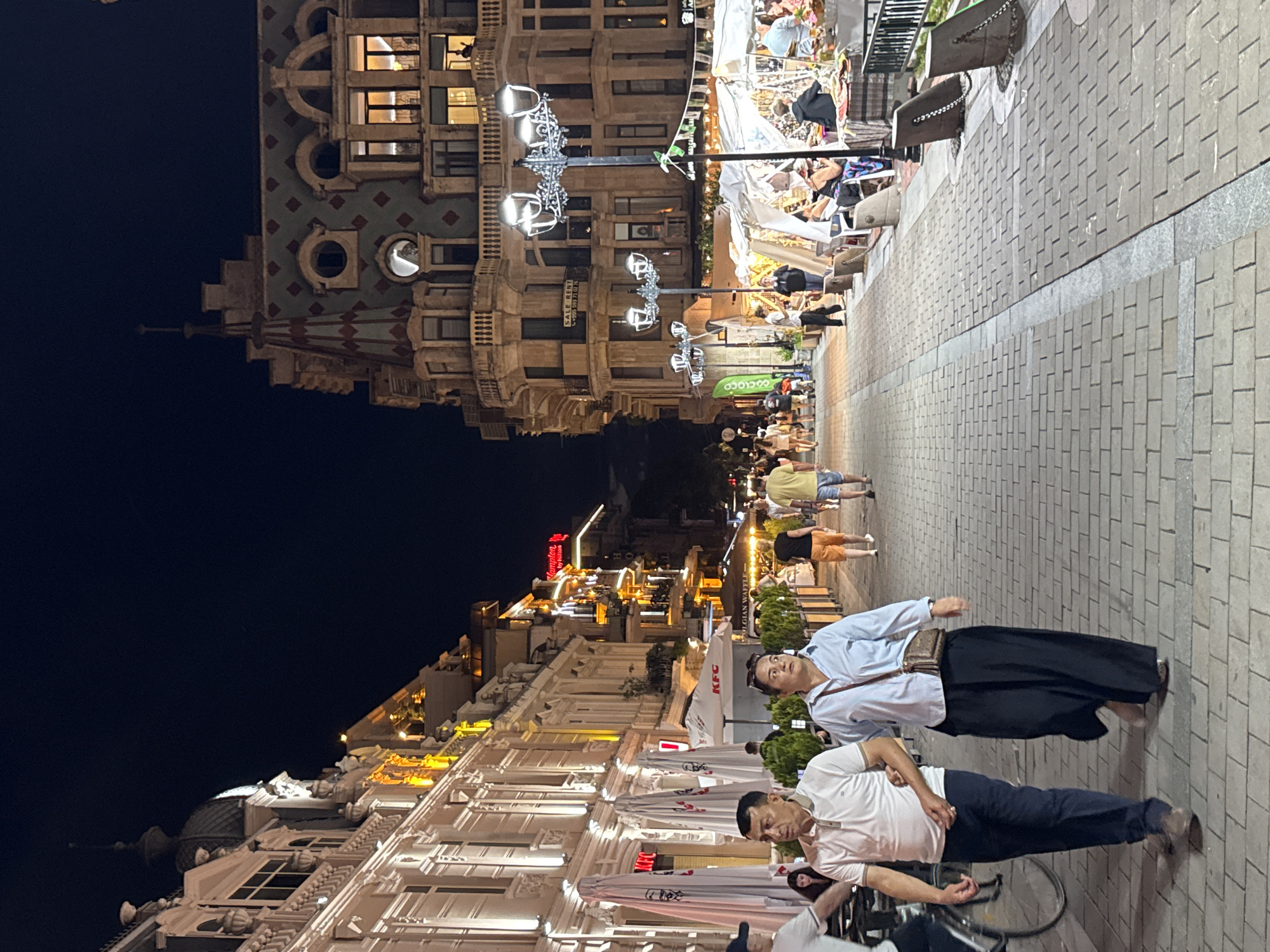 The image depicts a nighttime scene on a pedestrianized street in Batumi, Georgia, likely the Batumi Boulevard, a popular promenade. The street is paved with brick and lined with buildings and market stalls.
On the left, a multi-story building with ornate architecture is illuminated by warm yellow lights. Several umbrellas, some branded with "KFC," are set up outside what appears to be a restaurant. A man and a woman are walking towards the camera in the foreground. The man is wearing a white polo shirt and dark pants, with his left arm around the woman's waist. The woman is wearing a light blue shirt and dark, wide-legged pants, with a small crossbody bag.
On the right side of the street, another building with a distinctive patterned roof and arched windows is visible. In front of it, several market stalls are set up under white awnings, displaying various goods. People are browsing and interacting at these stalls. Street lamps with multiple lights illuminate the pathway. Further down the street, more buildings are lit up, and other pedestrians are visible. The overall atmosphere is one of a lively evening in a city center.