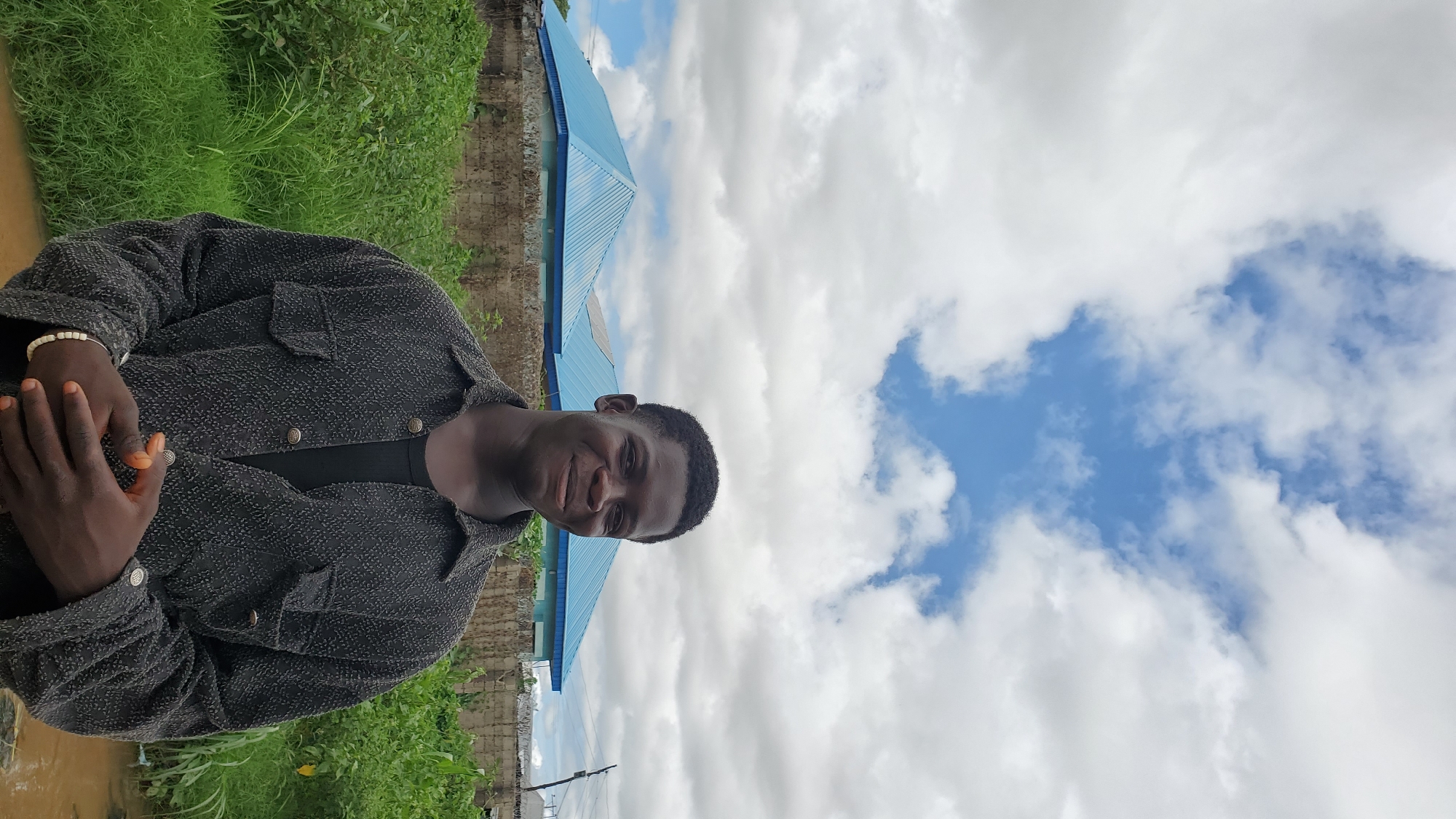 A young Black male stands outdoors, facing the camera with a slight smile. He is wearing a dark, textured button-down shirt over a black t-shirt, and his hands are clasped in front of him. A white beaded bracelet is visible on his left wrist. Behind him, a wall made of concrete blocks is visible, topped with a wire fence. Beyond the wall are two buildings with distinctive bright blue, sloping roofs. Lush green vegetation grows in front of the wall. The sky above is cloudy, with patches of blue peeking through. The scene appears to be in Jalingo, Nigeria, indicated by the architectural style of the buildings and the surrounding vegetation.