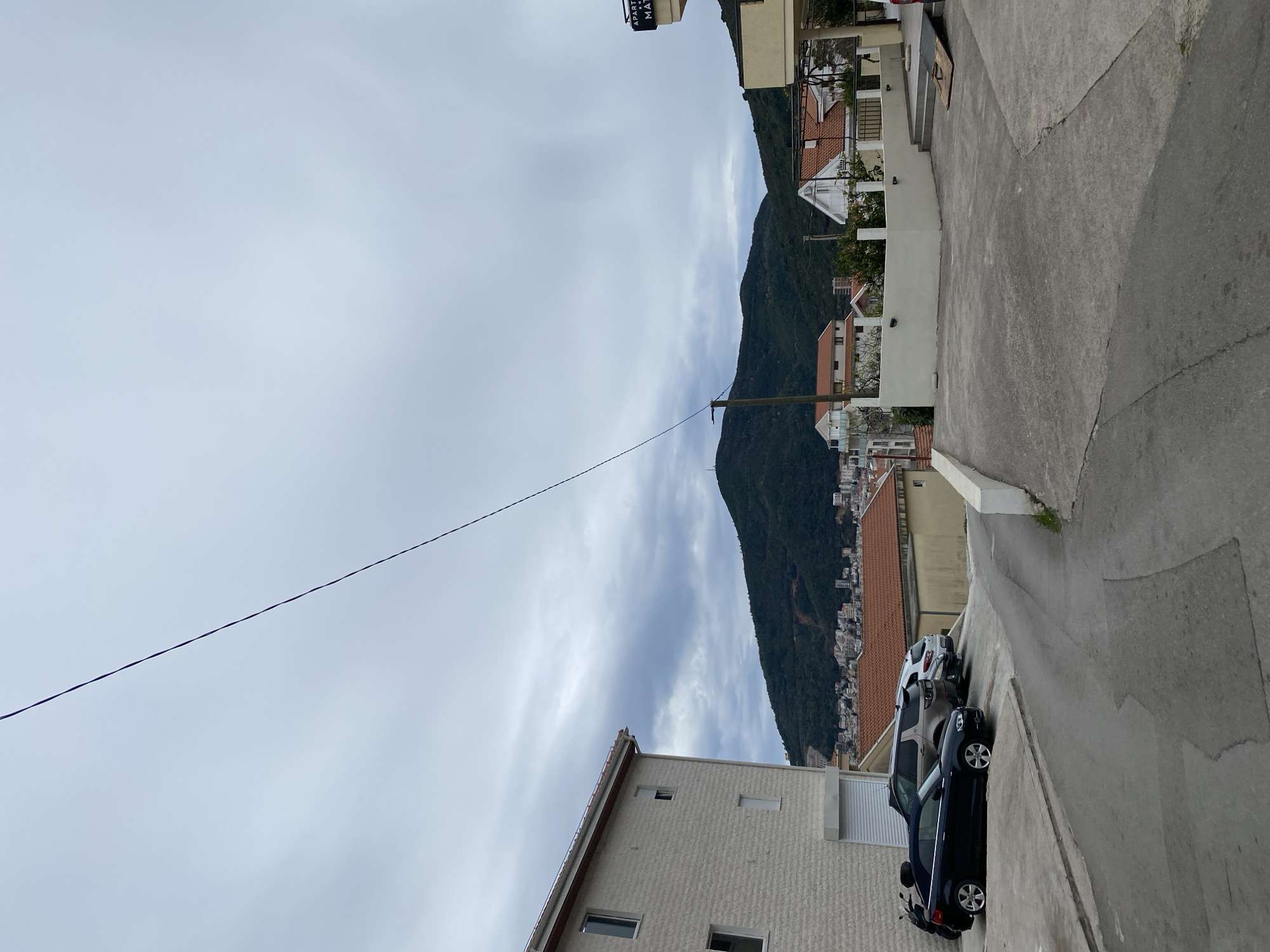 The image captures a street scene in Budva, Montenegro. A steep asphalt road curves downhill, lined with buildings on both sides. A lush, green mountain rises prominently in the background. The sky is overcast with a dense cloud cover. A single, taut power line stretches diagonally across the sky. Two parked cars are visible, a dark-colored sedan and a silver vehicle parked further down the road. The buildings along the street have a variety of architectural styles, with some featuring white or light-colored facades and red-tiled roofs.
