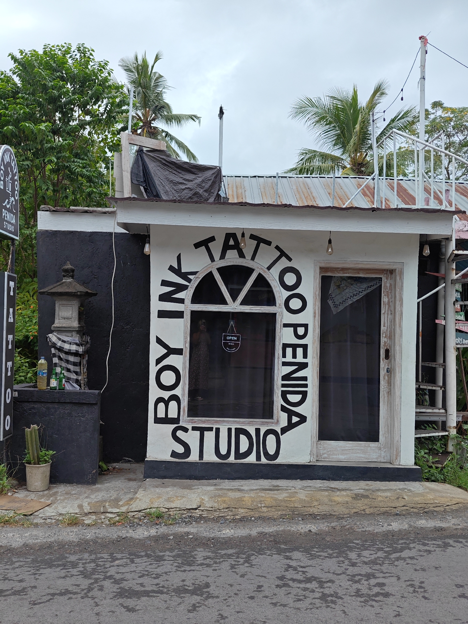The image captures the exterior of "Boy Ink Tattoo Penida Studio" in Nusa Penida, Indonesia. The studio is a small, single-story structure with a black and white facade. The name of the studio, "Boy Ink Tattoo Penida Studio," is prominently displayed in large black letters around the arched window and door. A stone lantern stands on a black base to the left of the entrance, with bottles and a checkered cloth placed on top. A potted cactus sits on the ground next to it. The roof is corrugated metal with a white trim. A black tarp covers part of the roof. A set of white metal stairs leads to the roof. A palm tree can be seen behind the building. The street is wet, indicating recent rain.