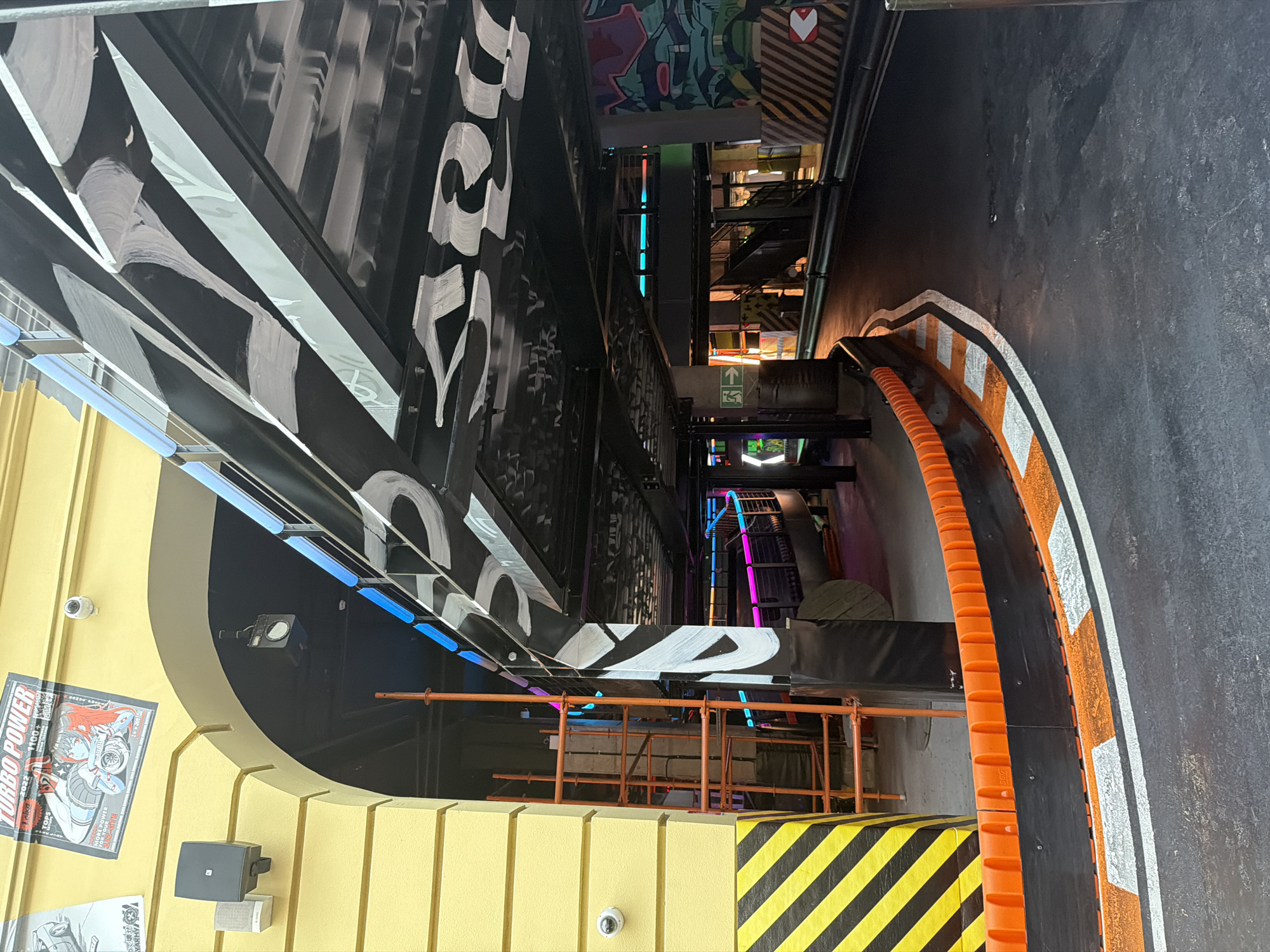This media file depicts an outdoor area that appears to be part of an entertainment or racing venue. The scene is dominated by large, black structures with white graffiti-style lettering and bold white stripes, suggesting an urban or industrial aesthetic. A bright yellow, curved architectural element is visible on the left, with posters attached to it. One poster shows an anime-style illustration and text that includes "TURBO POWER 2022" and "MORE POWER". A security camera is mounted on the yellow structure.
The ground is a dark asphalt surface with a painted track border, featuring orange and white markings, resembling a race track or go-karting area. There are also black and yellow hazard stripes visible on a low barrier. In the background, there are multi-level structures with scaffolding and stairs, illuminated by colorful neon lights, primarily blue and pink, adding to a vibrant and energetic atmosphere. The lighting suggests it could be evening or an indoor/shaded area designed to simulate nighttime. No people are clearly visible in the frame. The overall impression is one of a lively, dynamic environment, possibly a themed attraction or sporting facility.
