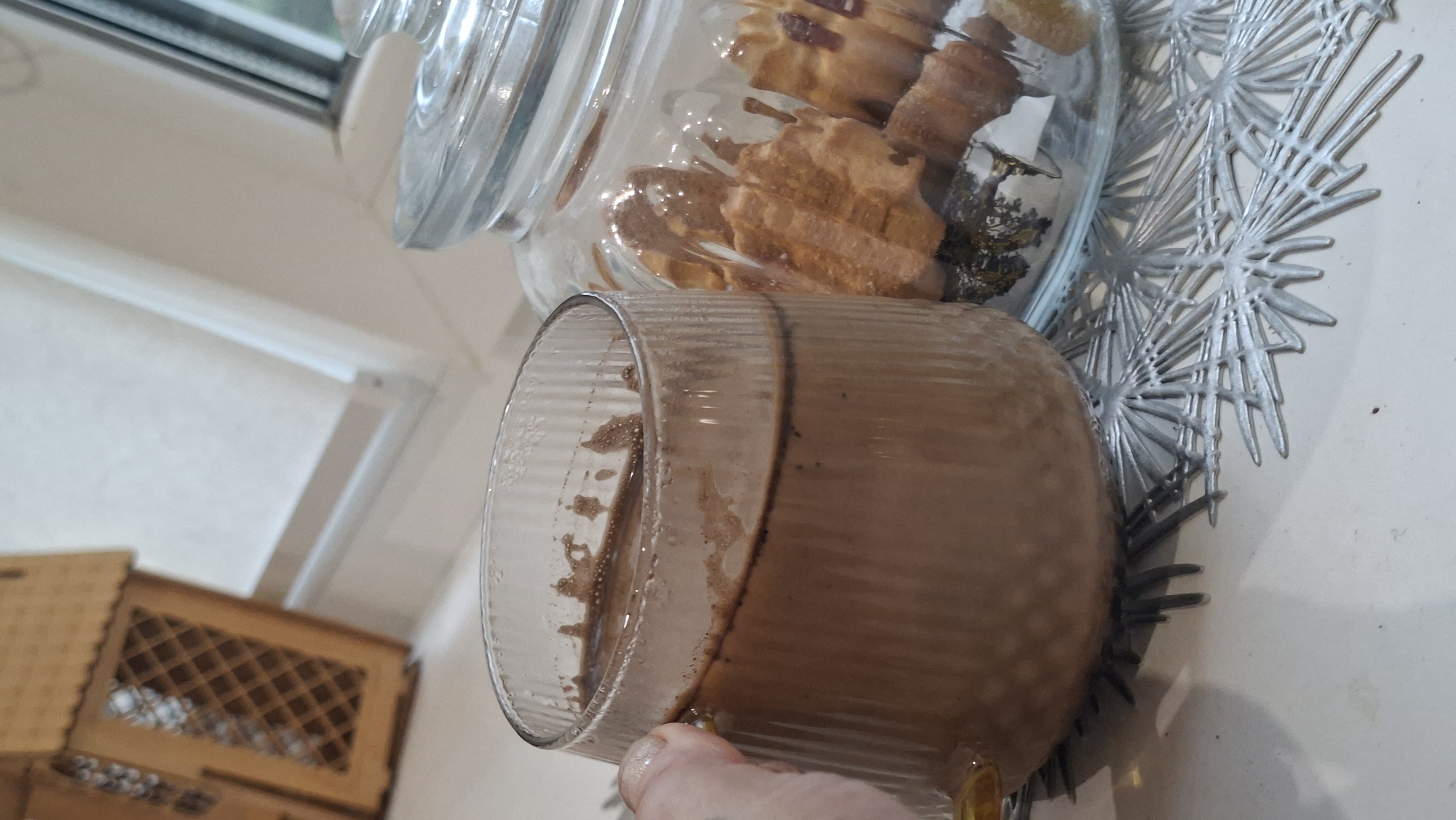 A person's hand is visible on the left, holding a clear, fluted glass mug filled with a brown beverage, likely hot chocolate or coffee. The mug is resting on a silver, star-shaped placemat. To the right of the mug is a clear glass jar with a lid, containing several cookies. The background shows a wooden decorative structure resembling a house on the left and a window with white blinds on the right. The overall scene appears to be indoors, on a countertop. The specific location in the city of Myadzyel, Belarus cannot be determined from the provided image.