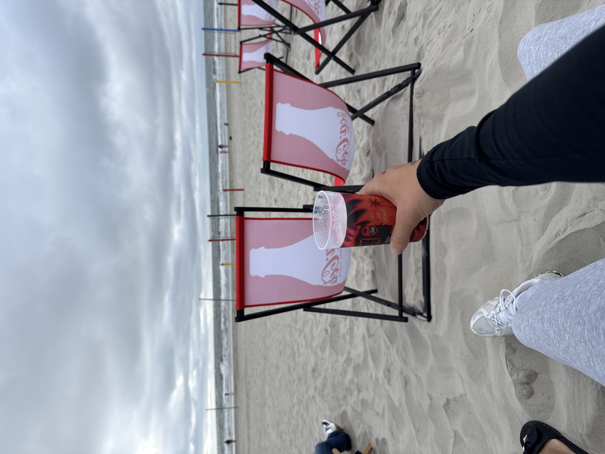 This image depicts a person holding a plastic cup of red liquid on a cloudy day at the beach in Langeoog, Germany. The cup has a label that reads "Beach Club" and shows palm trees and the words "SALT SAND SUNDOWNER". In the background, there are several red and white beach chairs with a Coca-Cola bottle logo printed on them. Beyond the chairs, the ocean and a grey, overcast sky are visible. The sand is textured with footprints and dunes. The person is wearing a black long-sleeved shirt and grey sweatpants. Another person's legs and feet, wearing white sneakers and black flip-flops, are visible in the lower left of the frame. Volleyball nets are set up in the distance, with a few figures walking along the shoreline.