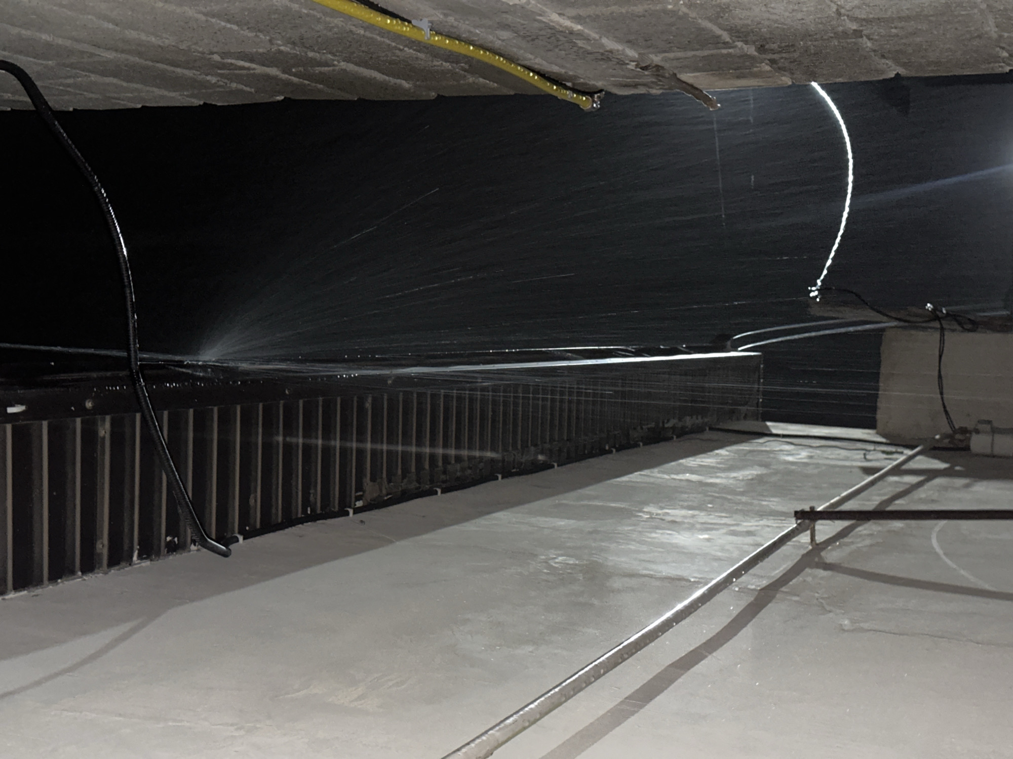 A narrow alleyway between two buildings is depicted at night. The building on the left has a smooth, light-colored wall with some dampness and streaking. A horizontal pipe or conduit runs along this wall, casting a shadow. A vertical metal pole supports this pipe in the foreground. The building on the right is constructed of rough, light-brown bricks and has a yellow cable attached to it with clips, the cable is also wet. A dark, corrugated pipe runs horizontally along the top of the opening between the buildings, attached to a slatted overhang. Water is spraying downward from this dark pipe, creating a curtain of droplets that catches the light. The background is dark, suggesting the absence of ambient light, with the water spray being the primary source of illumination within the frame. The scene appears to be in an outdoor urban environment, possibly a service alley or a passage between structures.