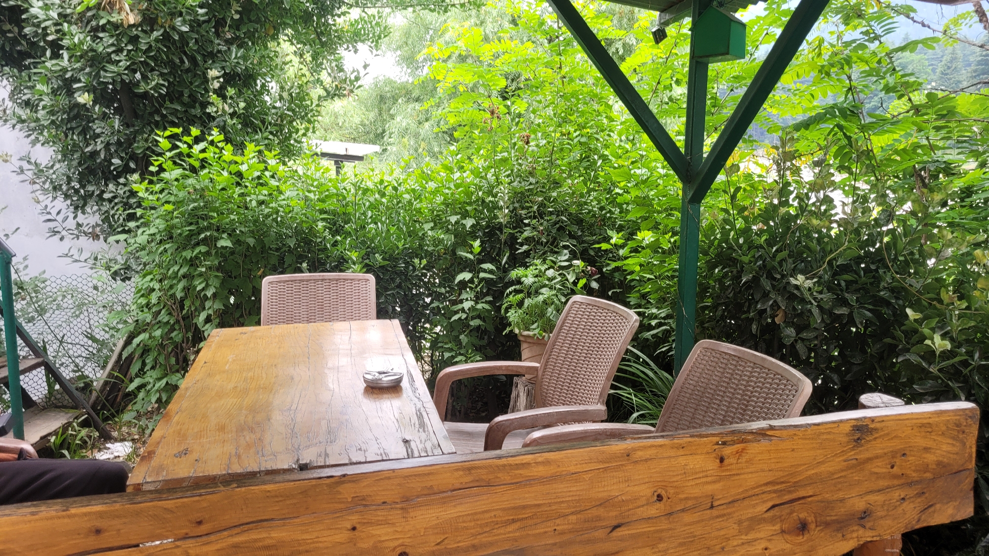 An outdoor seating area is depicted with a wooden table and chairs surrounded by lush greenery. The table, made of a light-colored wood with a visible grain pattern, is positioned in the foreground. On the table, there is an ashtray. Three plastic chairs with a woven texture are visible around the table. One chair is to the left of the table, another is behind it, and a third is to the right. A portion of a wooden bench is in the immediate foreground, obscuring the lower part of the scene. Behind the seating area, dense foliage creates a natural backdrop, with sunlight filtering through the leaves. A green metal structure, possibly part of a roof or pergola, is visible in the upper right quadrant of the image, with a small green box attached to it. The environment suggests a calm, possibly dining or resting space within a natural setting, consistent with the location of Vashisht, India, known for its hot springs and scenic beauty.