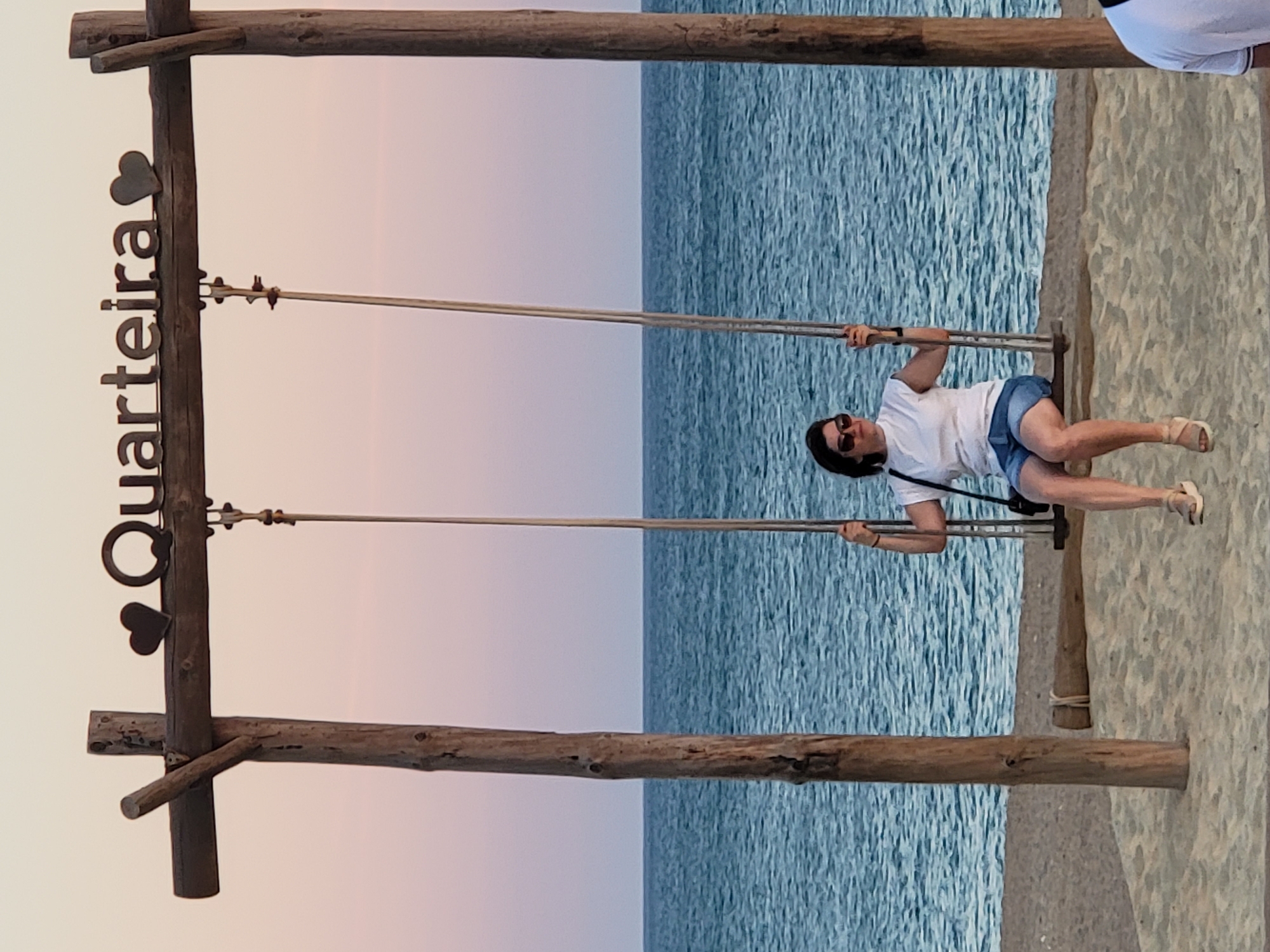 A woman with dark hair and sunglasses is sitting on a wooden swing on a sandy beach in Quarteira, Portugal. She is wearing a white t-shirt and blue denim shorts. The swing is suspended from a wooden frame that has the word "Quarteira" and two heart shapes attached to the top beam. The sea is visible behind her, with gentle waves and a clear sky. The lighting suggests it is either sunrise or sunset. The overall scene is peaceful and scenic, capturing a moment of relaxation by the ocean.