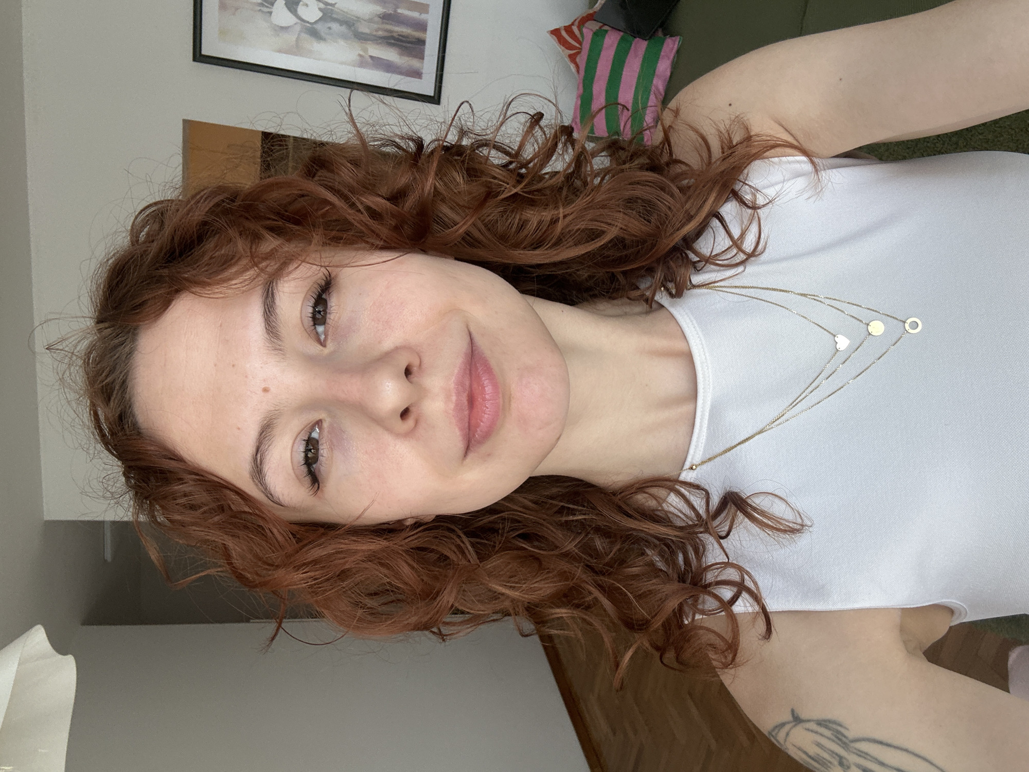 A young woman with fair skin, long, curly, reddish-brown hair, and dark eyes is depicted in a close-up, rotated selfie. She has a subtle, pleasant smile, looking directly at the camera. Her eyebrows are well-defined, and her eyelashes appear long. She wears a plain white sleeveless top and several delicate gold necklaces of varying lengths, adorned with small pendants including a heart, a solid circle, and an open circle. A faint, outline-style tattoo is visible on her left arm, peeking out from under her top.
The setting appears to be indoors, likely a home environment, with soft natural light suggesting it is daytime. In the background, there is a light-colored wall with a framed piece of abstract art featuring light, flowing colors within a dark frame. A pink and green striped pillow is visible, possibly on a sofa or bed. The image is cropped tightly around the woman, with elements of the room providing context rather than being a focal point. There is no visible text in the image, and while the context states Novi Sad, Serbia, there are no visual cues within the image itself to confirm this location. The overall impression is casual and relaxed.