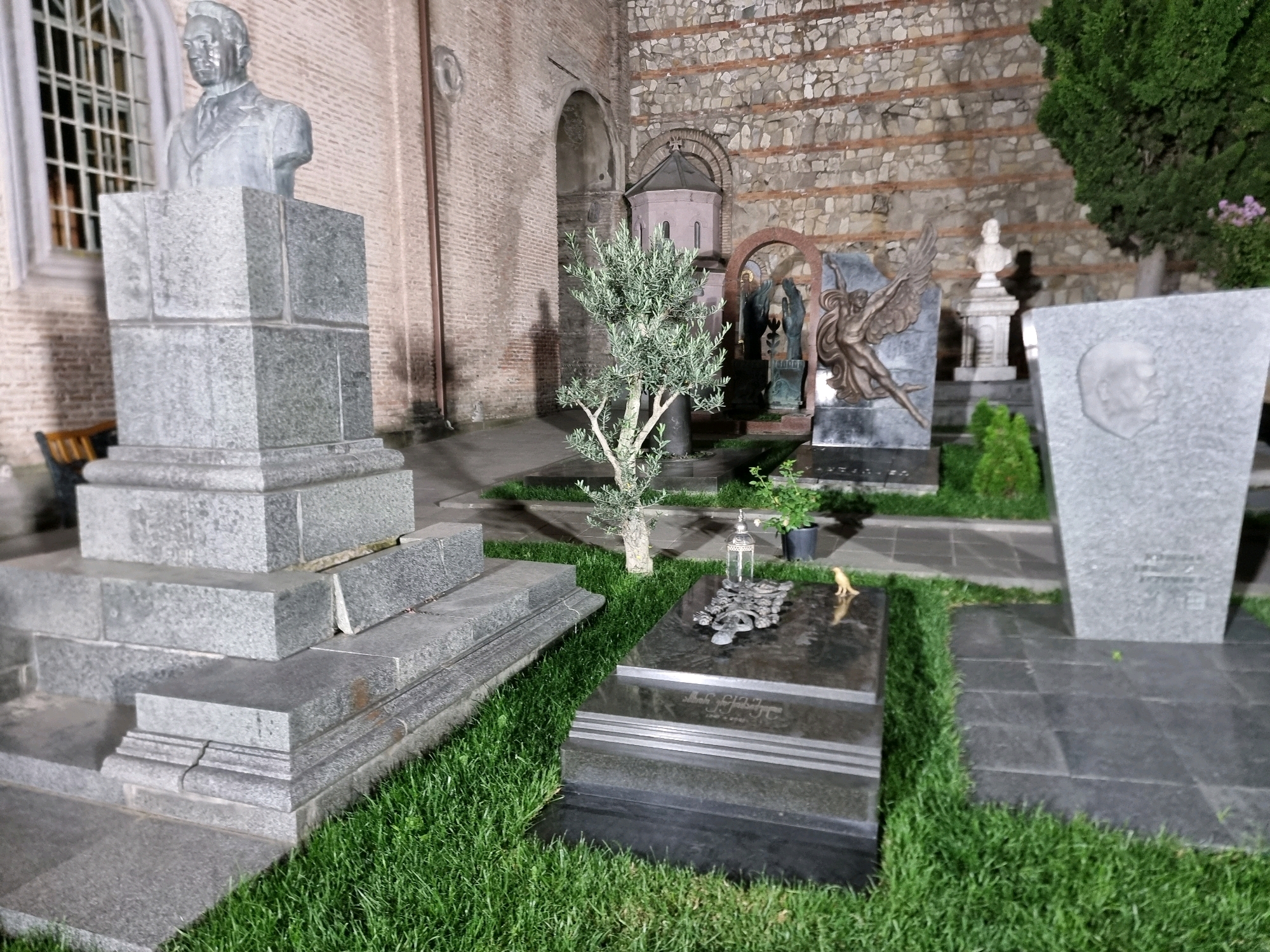 The image depicts a cemetery scene with multiple gravestones and statues. In the foreground, a granite bust of a man stands on a tiered pedestal. To the right, a dark granite tombstone with an inscription is partially visible, adorned with a metallic ornament and a small yellow bird figurine. Beyond that, a large, grey, angled headstone features a carved profile of a man's face. In the mid-ground, a young olive tree grows near a small, octagonal structure with a domed roof. Behind the tree, a sculpture of a winged figure is visible. The background shows a brick wall with stone accents, suggesting an older architectural style. The grounds are covered in well-maintained green grass and paved pathways. The lighting suggests it is dusk or nighttime, with artificial illumination highlighting the scene.