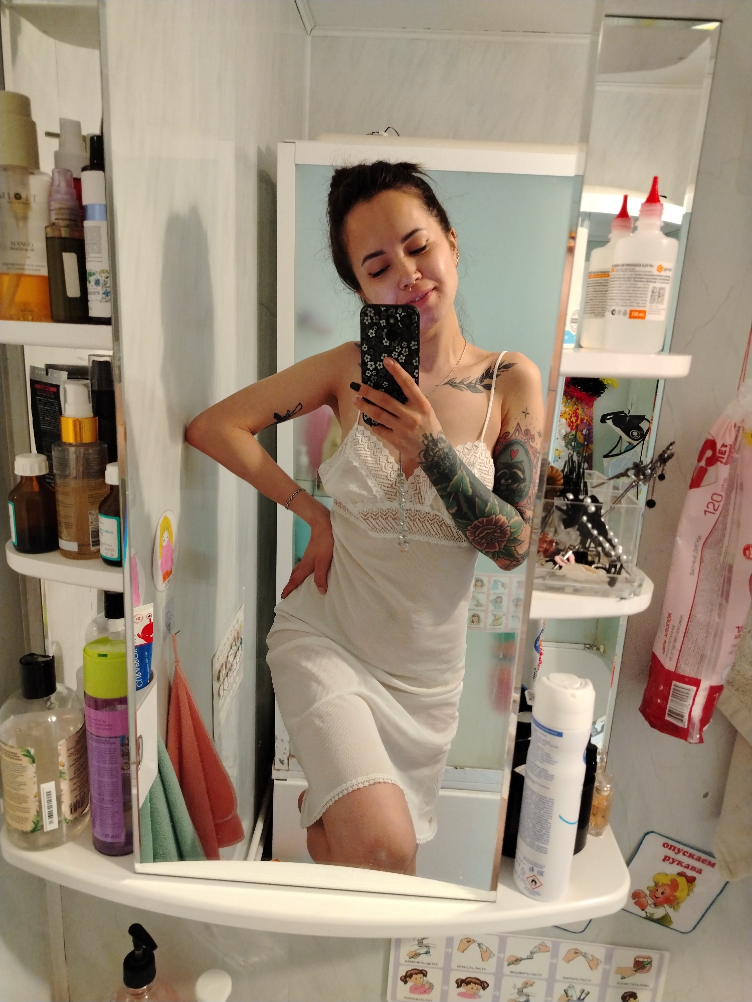 The image shows a young woman taking a selfie in a bathroom. She has dark hair pulled back in a bun, fair skin, and tattoos on her arms and chest. She is wearing a white slip dress. She is holding a smartphone with a floral case.
The bathroom is small and cluttered. There are shelves on either side of the mirror, filled with toiletries and cleaning supplies. There are also magnets and stickers on the side of the cabinet, as well as a card with instructions on proper hygiene.
The image may have been taken in the city of Noviy, Russia.