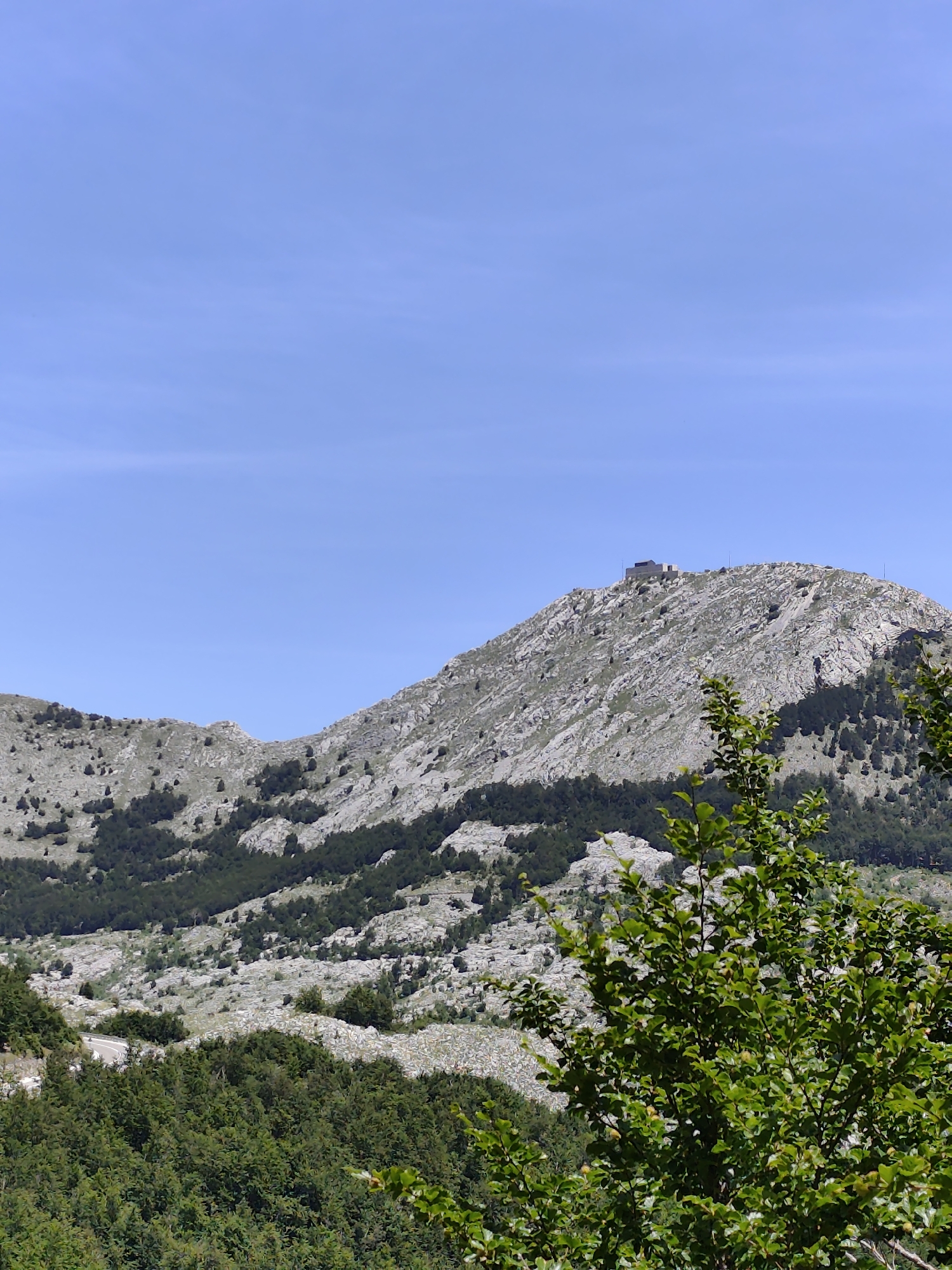 The image depicts a mountainous landscape with a clear blue sky. In the foreground, lush green foliage from trees partially obscures the view. The middle ground features a rugged mountain slope covered in sparse vegetation and rocky outcrops. Further up the slope, a cluster of trees forms a dark band against the lighter terrain. Dominating the peak of the mountain is a small, rectangular building with a flat roof, possibly a structure related to observation or communication, with several thin poles extending upwards from its vicinity. The overall scene suggests a remote, natural environment. The location is identified as Dolovi, Montenegro.