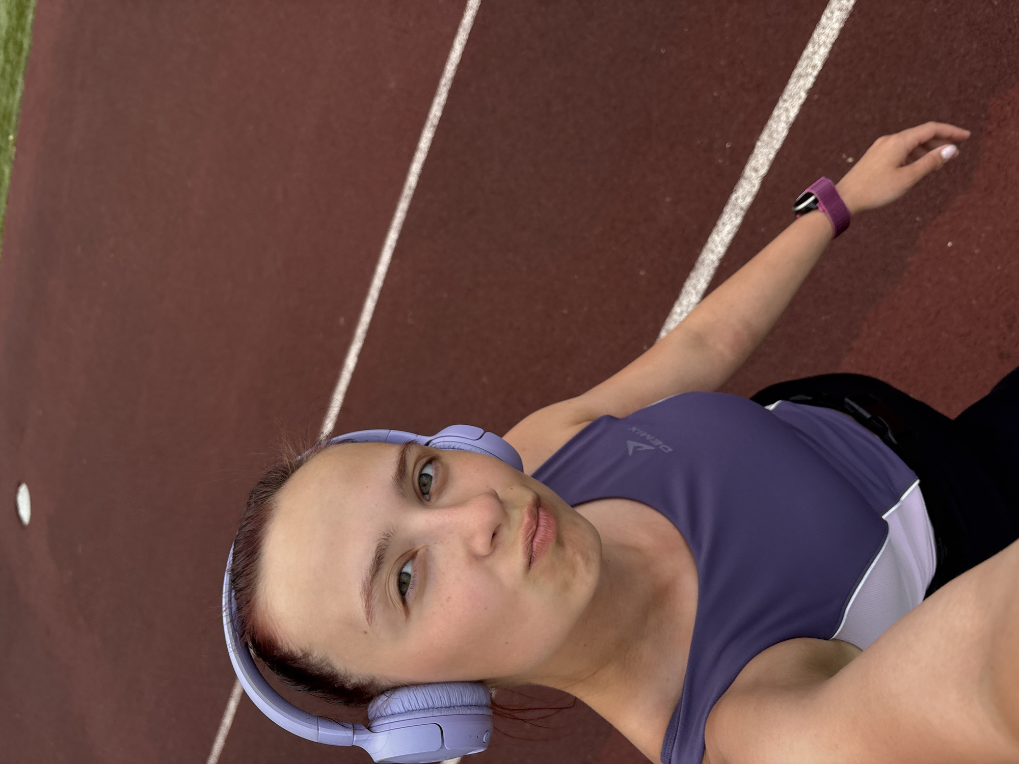The image shows a woman in Прикамский, Russia, presumably on a running track. She is wearing a purple tank top with a white stripe, black leggings, and purple headphones. She also has a purple watch on her left wrist. The track is maroon with white lines. There is a green area visible on the left side of the track. The photo is taken from a slightly high angle, likely as a selfie.