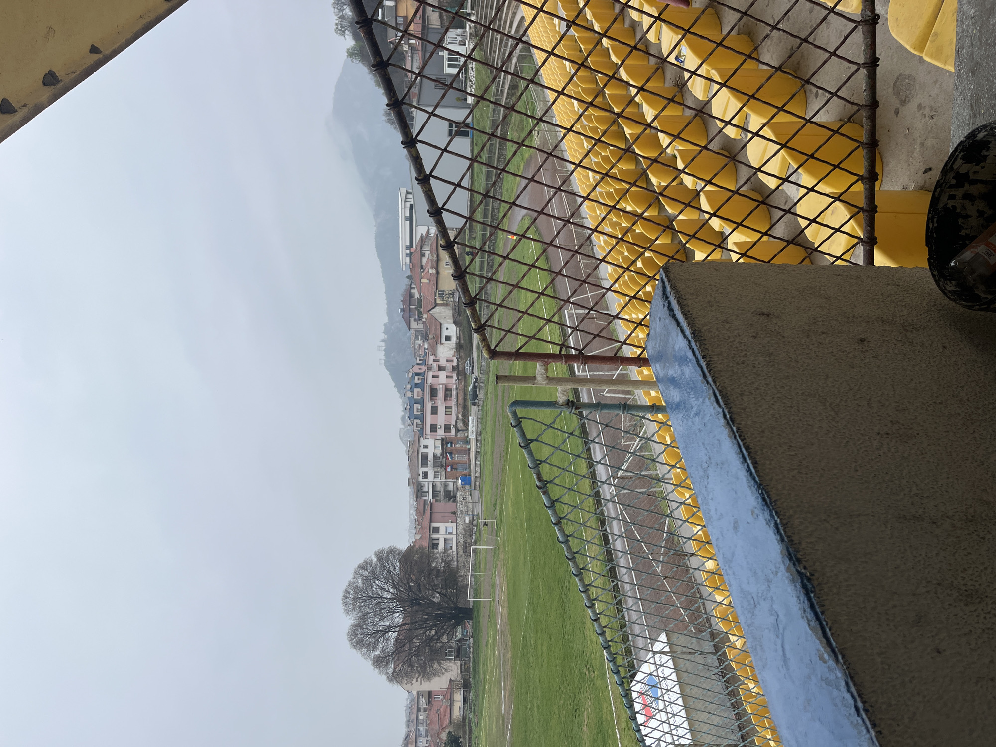 The image captures a view of the Asenovgrad Stadium in Bulgaria, seen from behind a chain-link fence and a concrete barrier. In the foreground are rows of yellow stadium seats. Beyond the fence, there is a grass soccer field with goalposts visible. Adjacent to the field is a running track, also enclosed by fencing. In the background, a residential area with buildings of varying heights and colors can be seen. In the far background, there is a mountain partially obscured by a cloudy sky. A bare tree stands near the edge of the field. The overall scene suggests a partly overcast day. There are no people visible in the image.