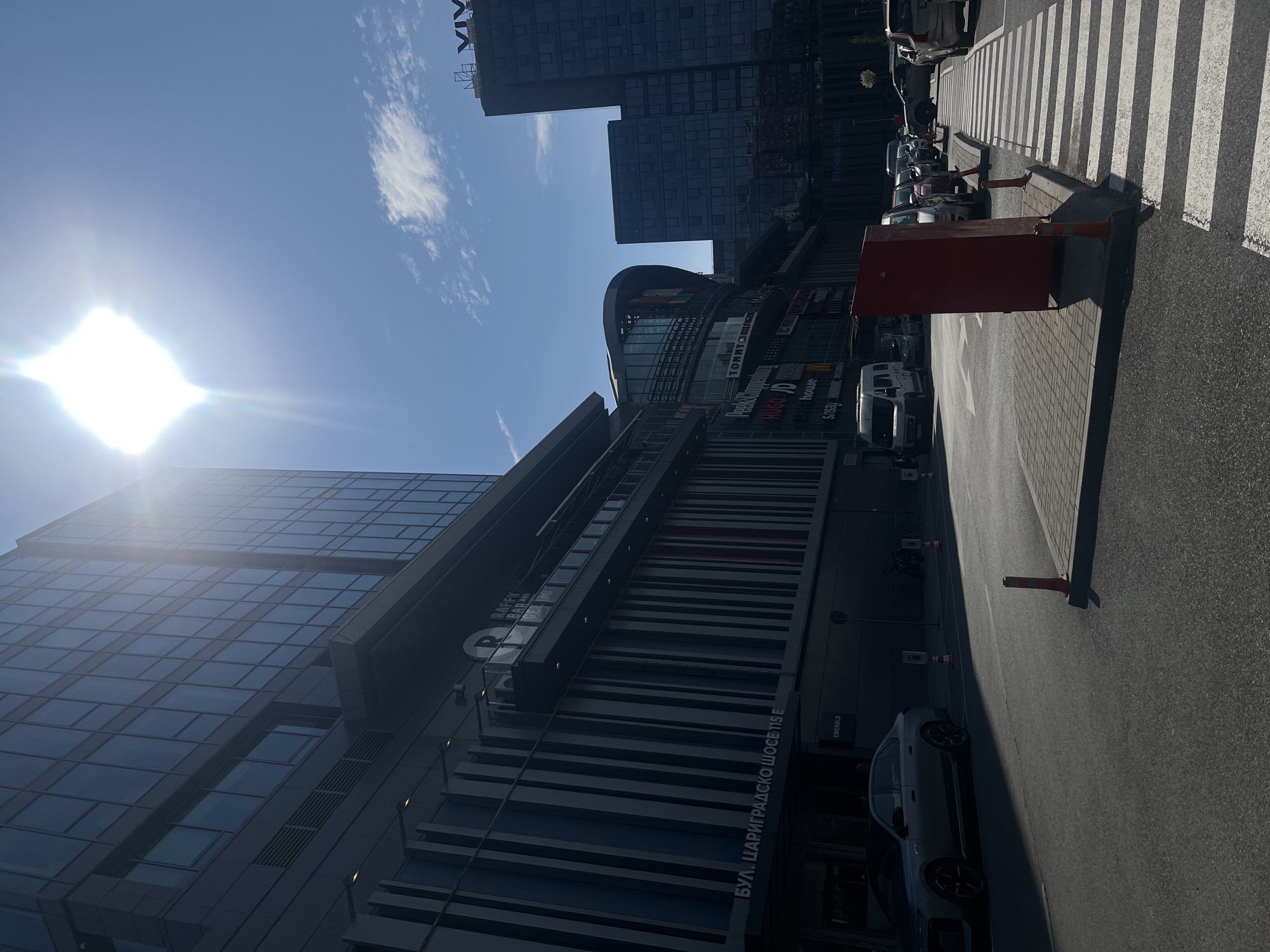 The image shows a street view in Sofia, Bulgaria, likely near the Paradise Center shopping mall. Modern buildings with glass facades and the VIVACOM headquarters tower are visible. Cars are parked along the street, and a crosswalk is present. Street furniture, including traffic barriers, is observed. The day appears sunny with a clear blue sky. The text "БУЛ. ЦАРИГРАДСКО ШОСЕ 115 Е" is inscribed on one of the buildings.