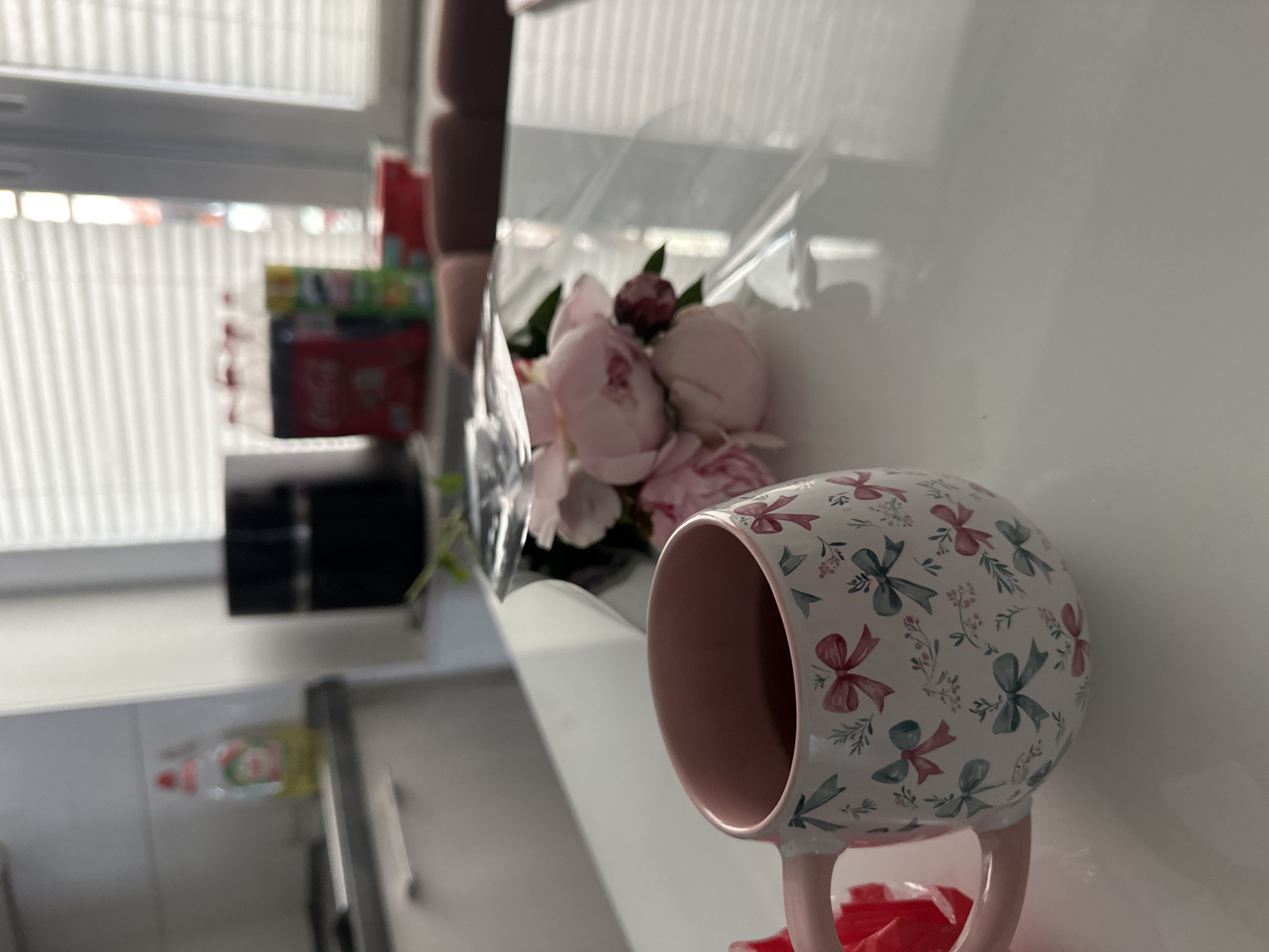 A close-up shot focuses on a white ceramic mug with a pink rim and handle, adorned with a pattern of pink and teal bows, sprigs, and berries. The mug appears to be filled with a dark liquid. To the right of the mug, a bouquet of pale pink peonies, some still in bud form, is wrapped in clear plastic. The scene is set on a reflective white surface, likely a table, which mirrors the objects on it. In the background, a kitchen counter is visible with a bottle of dish soap and a black appliance. Further back, through a window with horizontal blinds, a glimpse of the outdoors or another room can be seen. The overall lighting suggests a bright day, possibly indoors in a home or cafe. The image lacks any specific indicators placing it in Kutenholz, Germany, or any other specific city.