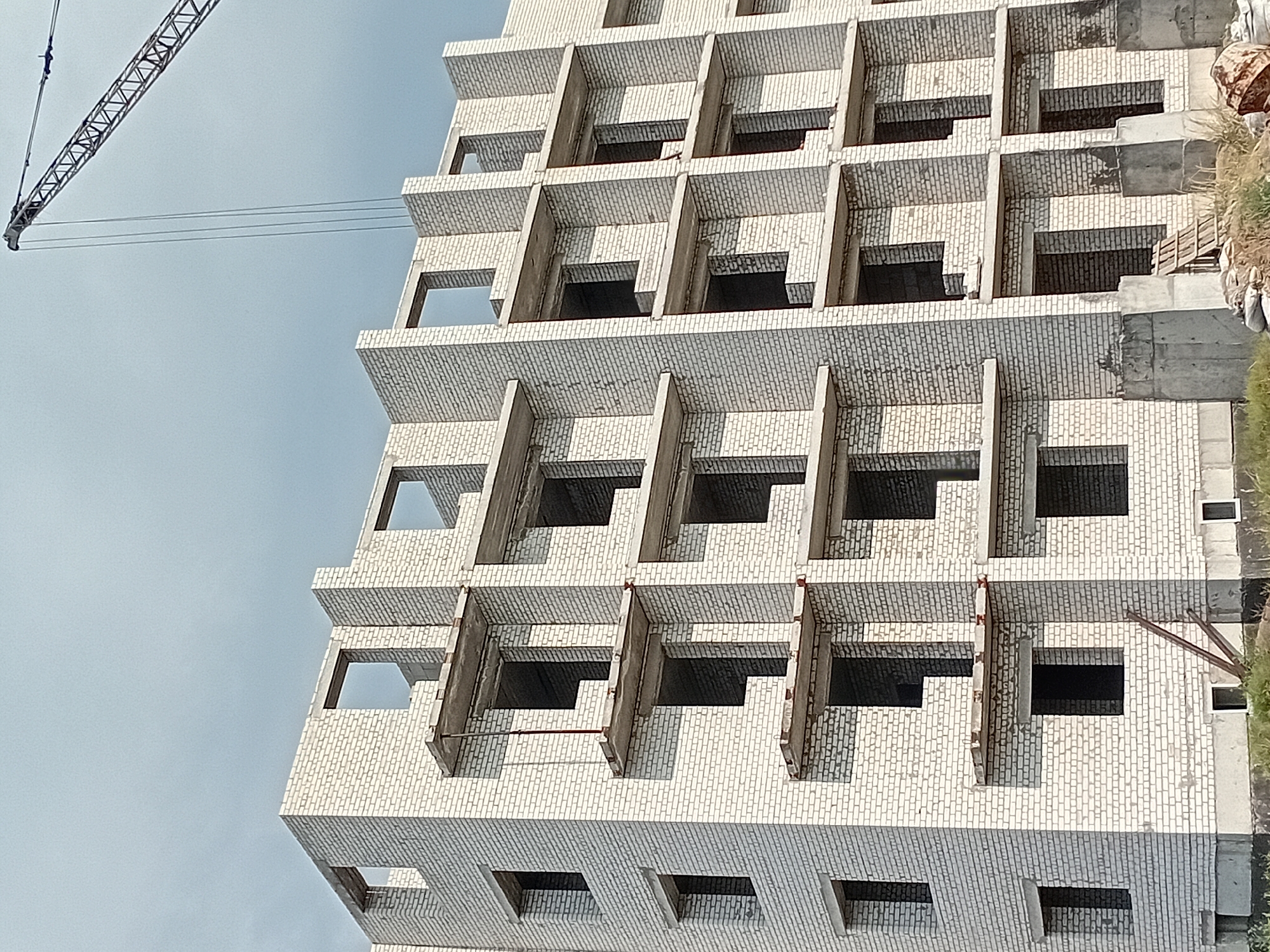 The image captures a multi-story building under construction, made of white brick, in the city of Semey, Kazakhstan. The building has numerous window openings and protruding concrete ledges on each floor, suggesting it is designed for balconies or structural support. A construction crane is visible in the upper right corner, with its arm extending over the building. The sky is overcast with a cloudy, gray appearance. In the foreground, there are piles of construction materials including sandbags and what appears to be wooden scaffolding or planks. The ground is covered with dry grass and some dirt. The scene depicts an active construction site, indicating ongoing development or renovation.