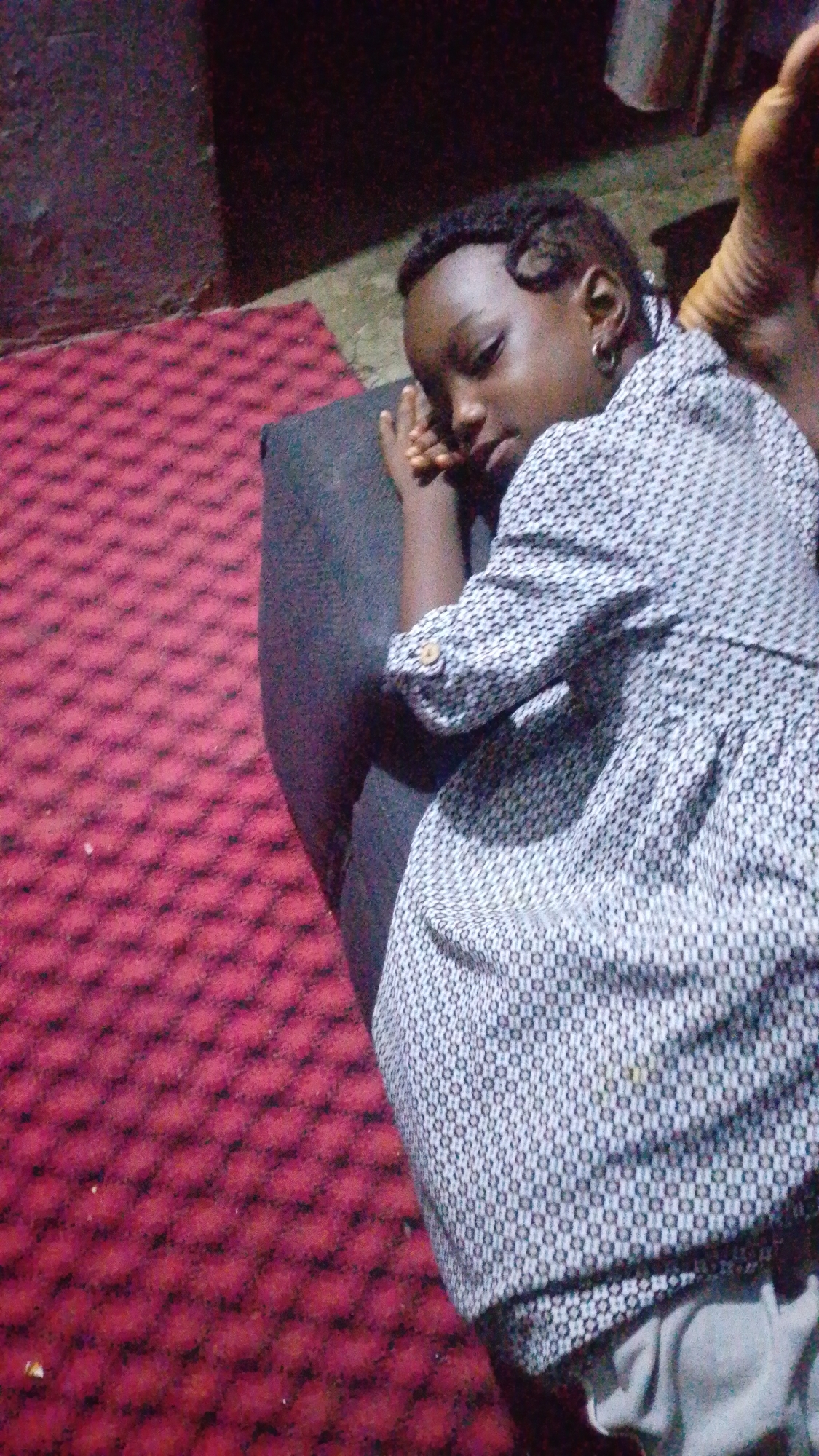 A young Black person with braided hair and a hoop earring lies on their side, sleeping. They are wearing a patterned shirt and grey pants. The person is lying on a dark grey cushion next to a red patterned carpet. The background is dark and out of focus. The scene appears to be indoors in an area of Unknown, Unknown.