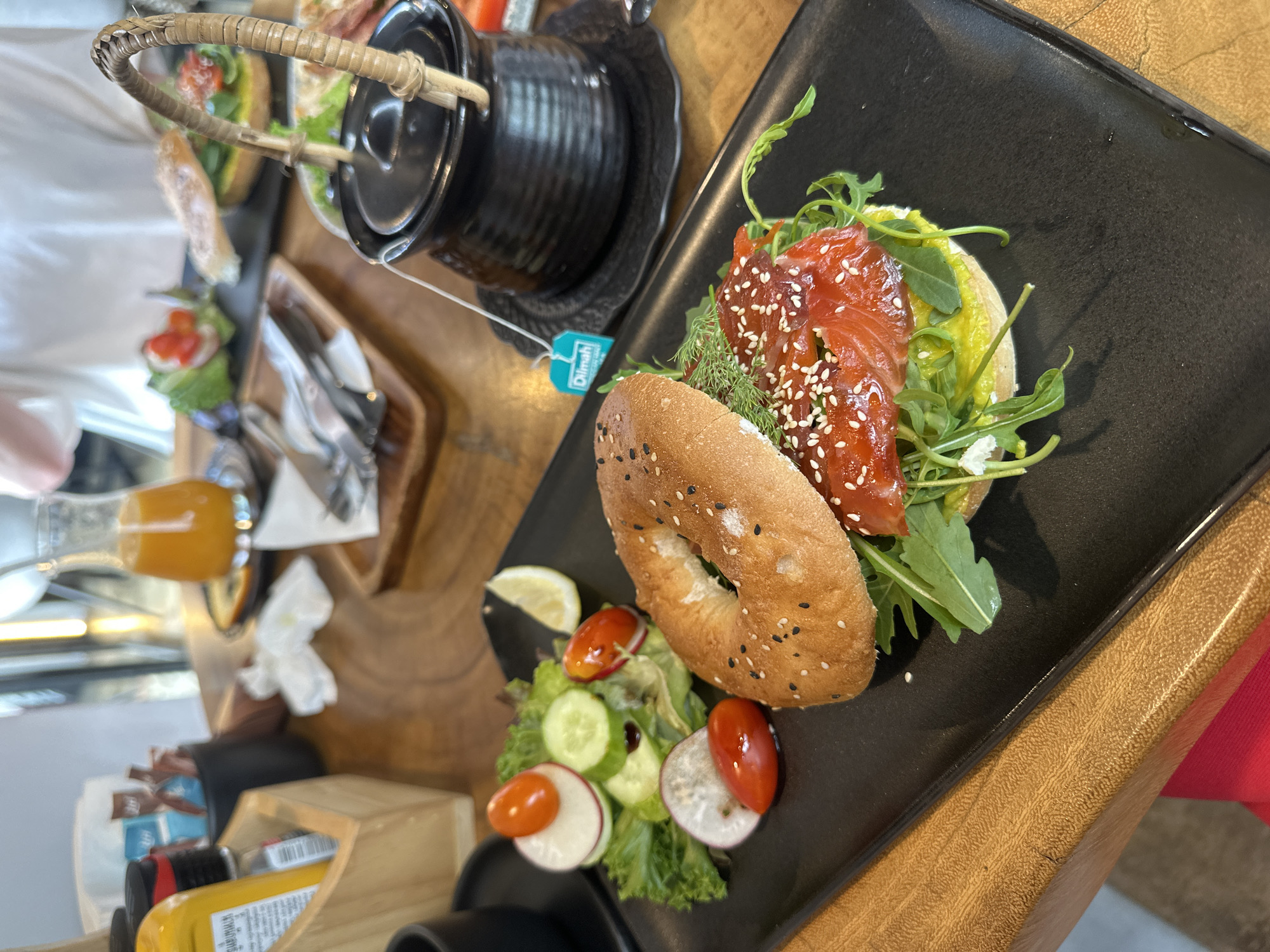 A bagel sandwich sits on a black rectangular plate alongside a small salad of lettuce, sliced cucumbers, radishes, and cherry tomatoes. The sandwich consists of a sesame seed bagel, leafy greens, and a stack of salmon with sesame seeds on top. Next to the plate is a small black tea pot with a bamboo handle. A glass of orange juice and silverware sit on a wooden tray to the left of the plate. Also visible are condiments, a wooden organizer, and other plates with food. The image was likely taken in Baan Bo Phut, Thailand.