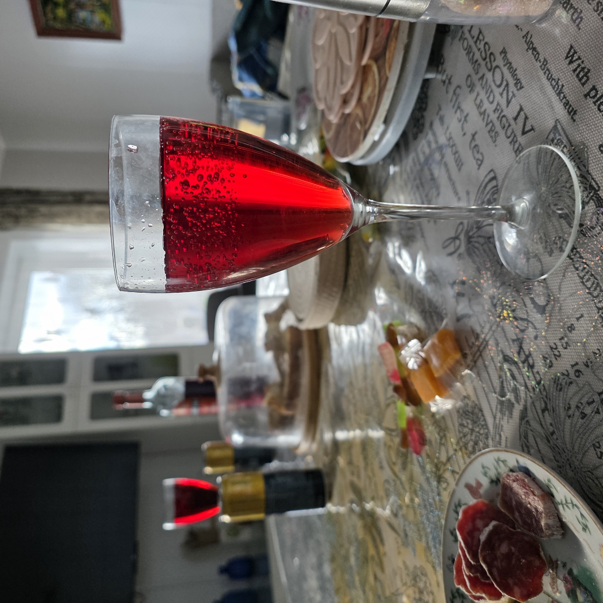The image depicts a table setting with a glass of red liquid, possibly wine, prominently displayed in the foreground. The table is covered with a printed tablecloth featuring text and botanical illustrations.
Additional food items are present, including a plate of sliced sausage and a platter with what appears to be dried fruit.
The environment includes a window in the background, suggesting an indoor setting. The presence of furniture further indicates the scene is inside a home or establishment.
The location is likely Koshelevichi, Russia, inferred from the context of the image origin. No people are visible in the scene. The objects are arranged for a meal or gathering.