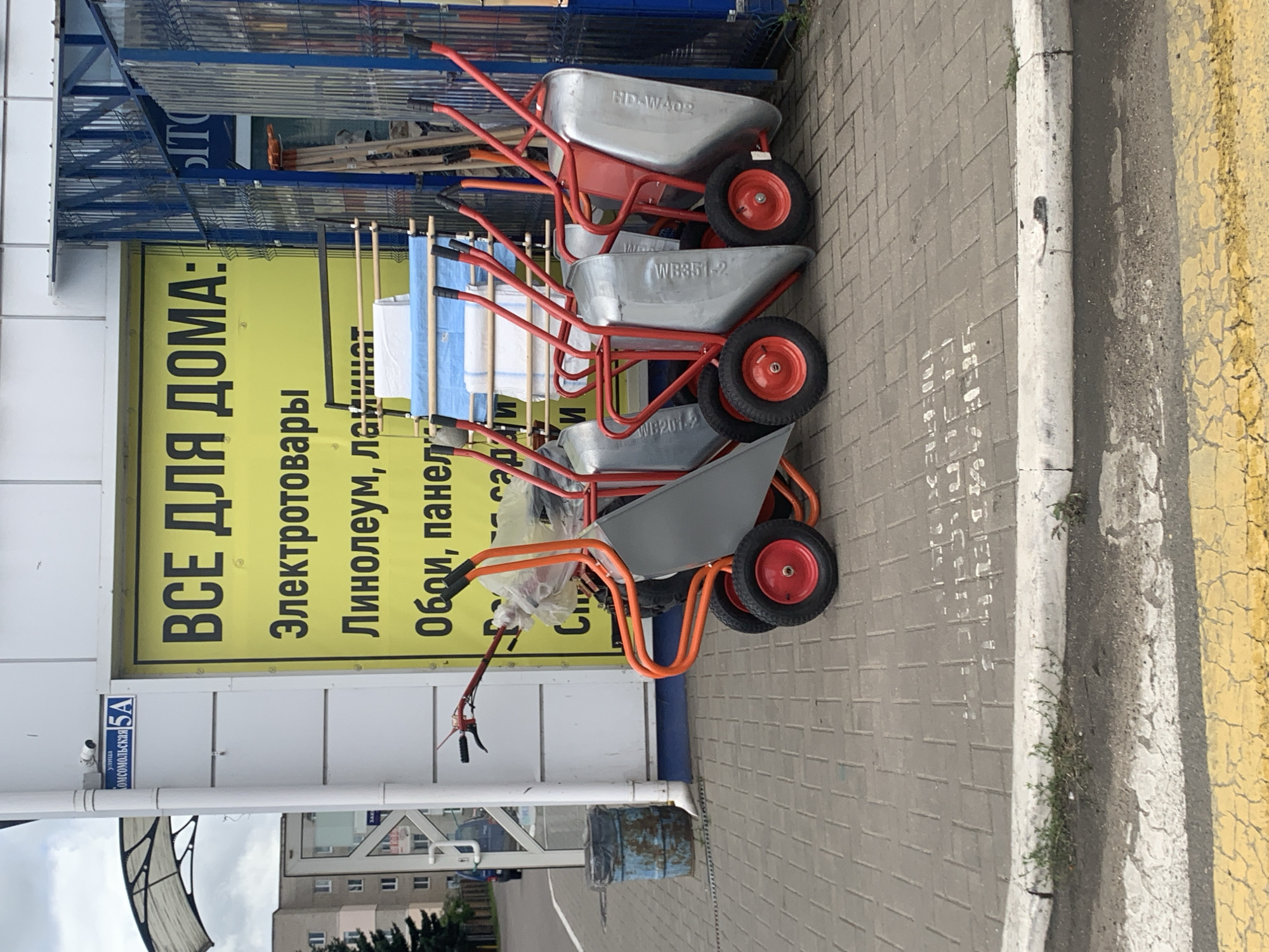 The image captures a row of red and silver wheelbarrows lined up on a paved sidewalk in front of a building. A large yellow sign with black text, which appears to be an advertisement, is visible behind the wheelbarrows. To the left of the sign, a white pole with a blue street sign reading "ул. Комсомольская 5А" is attached to the building. The building facade is white and appears to be made of metal panels. A blue dumpster with a rusted metal band is situated to the left of the wheelbarrows. The ground is a brick-patterned pavement with a white and yellow painted curb and road markings in the foreground. The sky is overcast. The scene is outdoors, likely in a commercial area. The location is identified as Kapyĺ, Belarus.