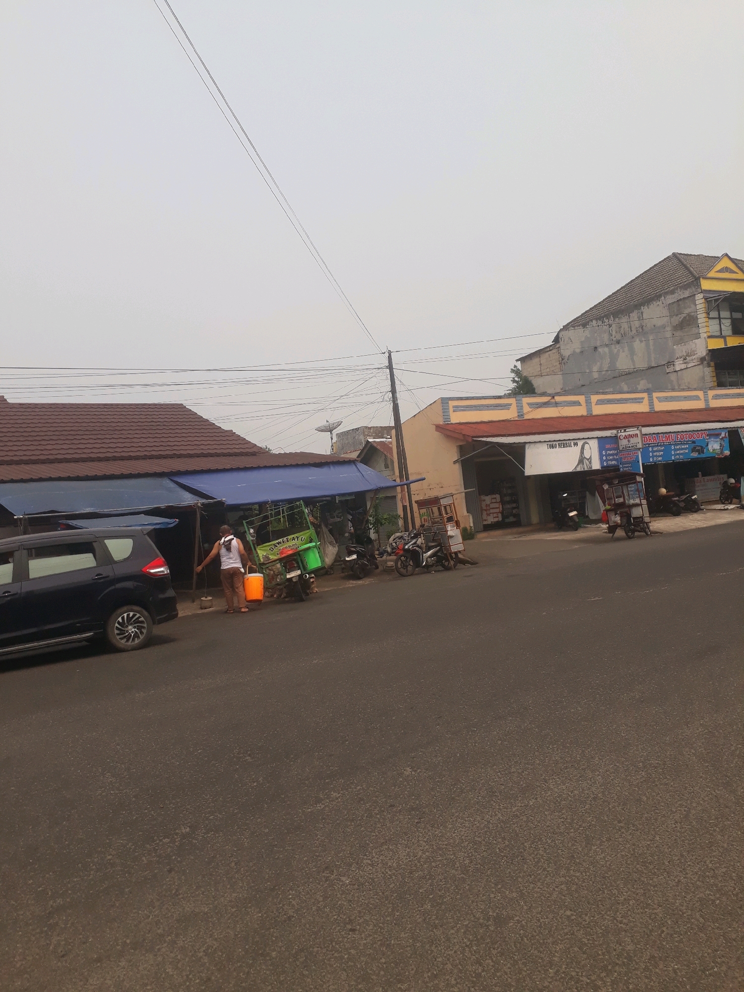 The image depicts a street scene in Pangkalpinang, Indonesia, characterized by a hazy, overcast sky. In the foreground, a paved road is visible, with a dark blue minivan parked on the left. A person wearing a white tank top and brown pants is standing on the sidewalk next to a green motorized tricycle, holding a bucket and a broom. Behind the person, a building with a brown tiled roof and blue awnings lines the street. Further down the road, another building with a yellow and beige facade displays signage that includes "Canon" and "DRY ILMU FOTCOPY," suggesting a business. Several motorcycles are parked in front of these businesses. The overall environment suggests a commercial area in a residential setting.