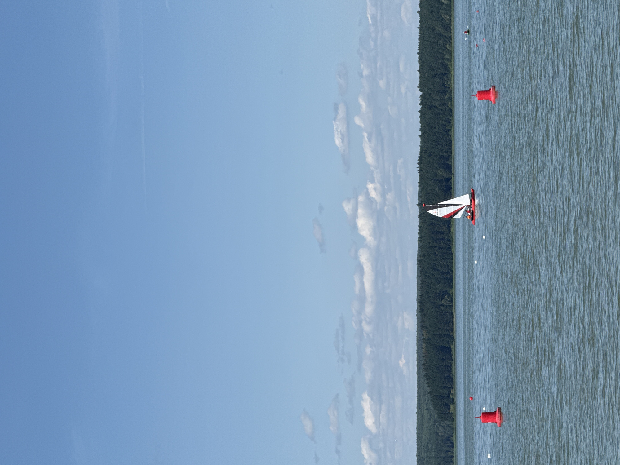 A red and white sailboat with two individuals on board sails across a calm body of water. In the foreground, two red buoys mark a section of the water. A dense line of dark green trees forms the horizon, with a clear blue sky above dotted with scattered white clouds. The scene appears to be the Votkinsk Reservoir in Votkinsk, Russia, a popular recreational area known for its water activities.