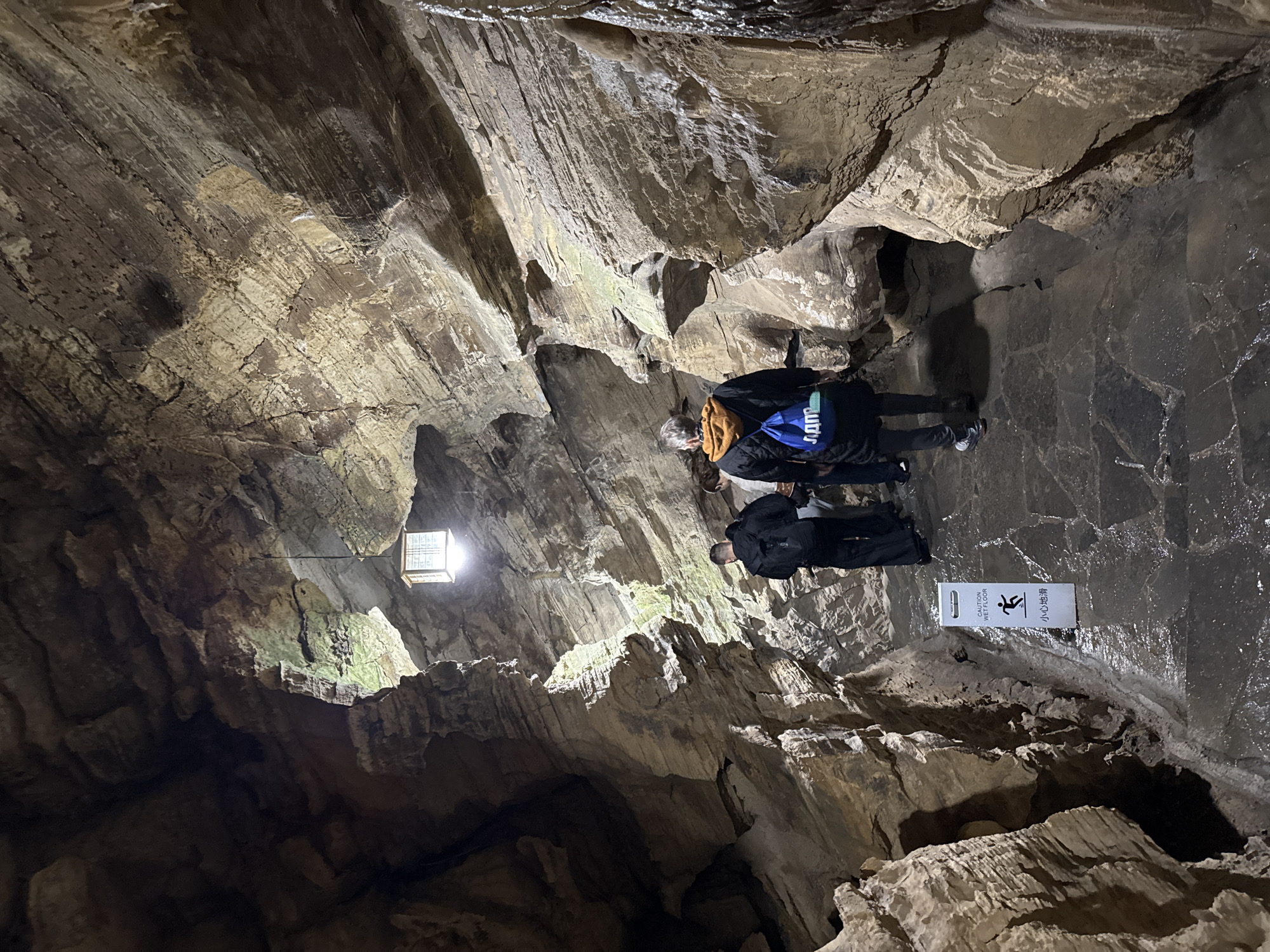 The image depicts the interior of a cave in Wulong District, China. Two individuals are present. One wears a black jacket with a blue backpack with yellow drawstring, while the other wears a black jacket. A sign is visible, with an image of a stick figure sliding on a wet surface, and the words "CAUTION WET FLOOR," in English and Chinese. The cave walls are textured and illuminated by a rectangular light fixture. The floor appears to be wet stone.