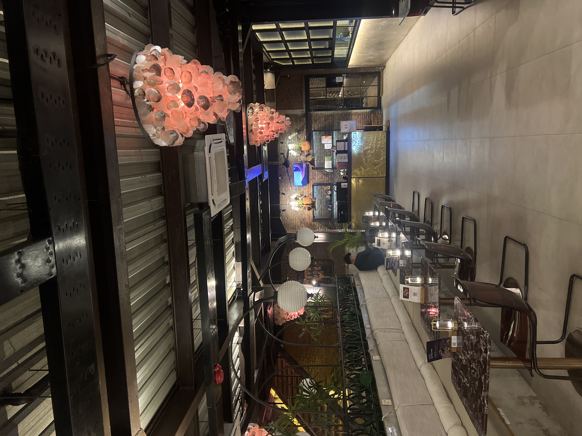 The image shows a multi-level interior space in North Kuta, Indonesia. The architecture features exposed metal beams and corrugated metal panels on the ceiling, giving an industrial feel. Decorative lighting fixtures, some resembling coral and others spherical, add ambiance. A long, light-colored sofa lines one side, with small tables and seating along a raised bar. Plants are integrated into the decor. A person is seen sitting on the sofa. The space seems to be a bar or restaurant, with a mix of materials.