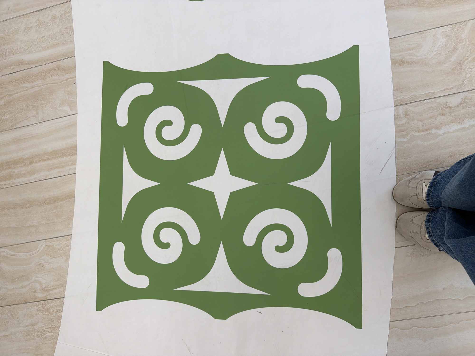 The image shows a white sheet with a green stencil pattern on it. The pattern is symmetrical and features four swirling shapes arranged around a central star. The stencil is placed on a light-colored tiled floor, and a pair of feet wearing jeans and sneakers are visible at the bottom of the frame. The scene is an indoor environment, likely a home or studio, and there is no indication of Yuzhno-Sakhalinsk, Russia.