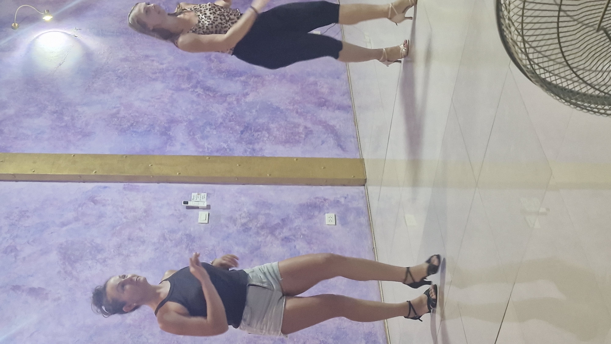 The image shows two women on a tiled floor in front of a purple-hued wall. The lighting is artificial and bright, suggesting an indoor setting, possibly a studio or a dance floor. The wall has a textured, mottled appearance, predominantly in shades of purple with some lighter marbling. A horizontal golden beam or shelf runs across the middle of the image.
In the upper portion of the image, a woman is seen from the waist down. She is wearing a leopard-print top and black capri pants, along with gold-strapped high heels. Her legs are positioned as if she is in mid-movement or posing.
In the lower portion of the image, another woman is visible, also from the waist down. She is wearing a black tank top and light-colored denim shorts, paired with black strappy high heels. Her arms are slightly raised, suggesting she is also engaged in a dance or movement. The floor is tiled with large, pale squares, and a reflection of the scene is visible on the polished surface. To the right of the frame, a portion of a large, circular fan with a wire cage is visible.
The overall impression is that of an active or performance-oriented environment, with both individuals seemingly in the midst of dancing or practicing. The reflections on the floor add a sense of depth and dynamism to the scene. There is no visible text in the image.