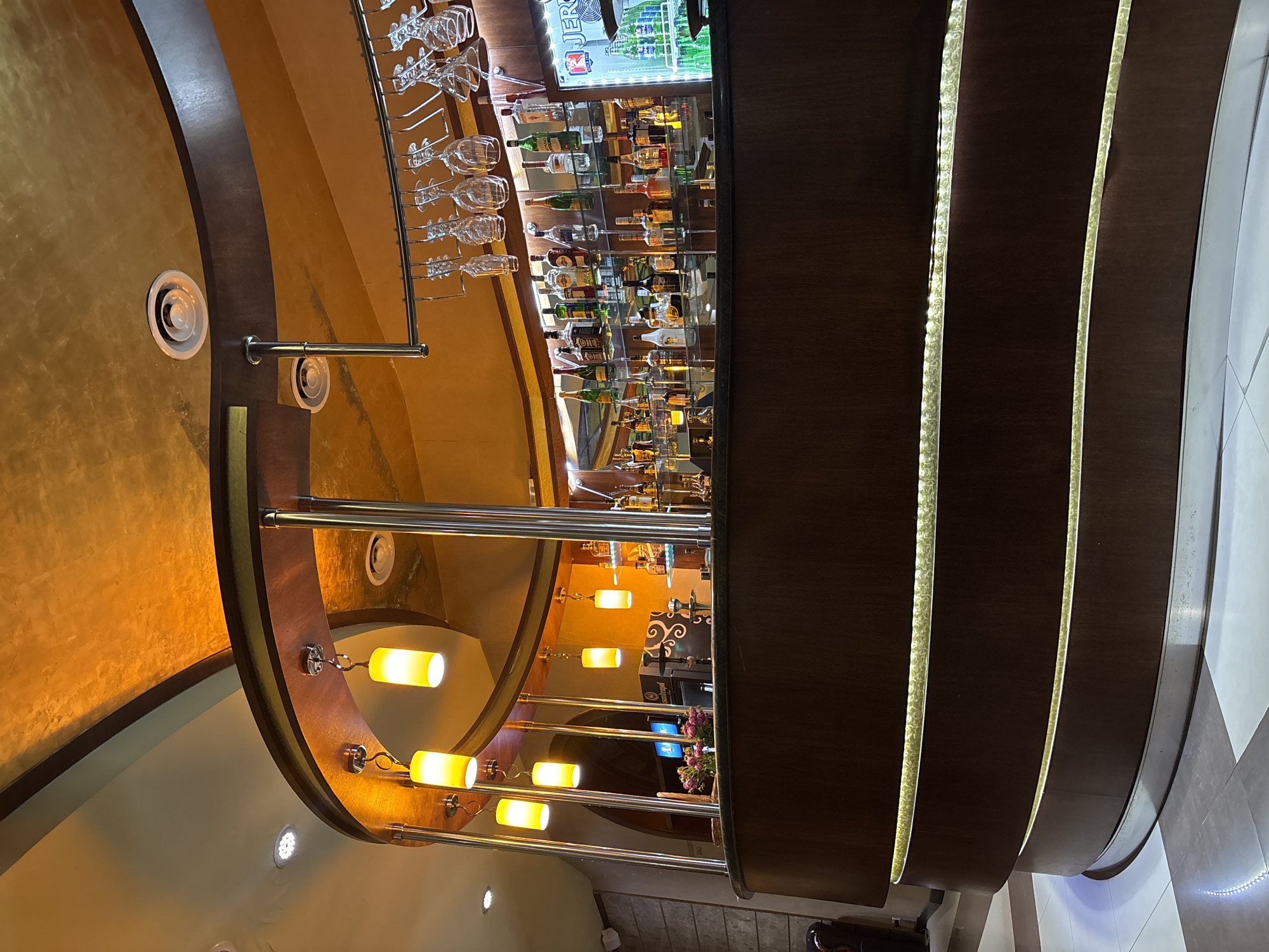 The image shows an interior view of a bar area. The bar itself is made of dark wood with curved, layered sections, illuminated by soft golden lights embedded in the curves. Above the bar, numerous bottles of liquor are displayed on shelves behind a mirror. Several wine glasses are hanging upside down from a rack above the liquor bottles. The ceiling is a textured golden color and features recessed lighting and air vents. Several cylindrical pendant lights with yellow illumination hang from the ceiling, suspended by metal rods. In the background, there appears to be a television screen displaying an image and some decorative floral arrangements on the bar counter. The overall impression is of a well-lit, sophisticated bar, possibly in a restaurant or lounge. There are no people visible in the image.