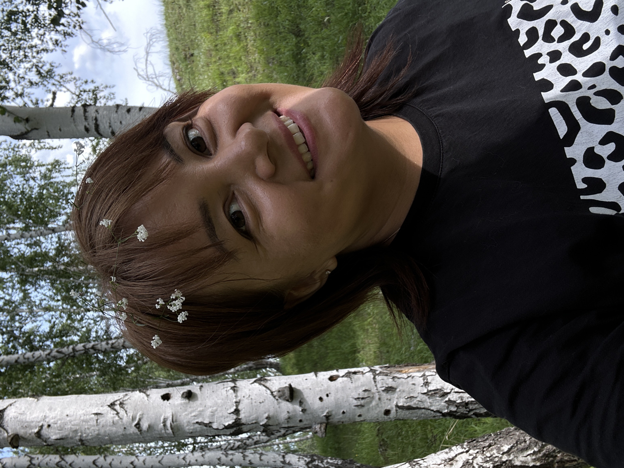 A young woman with brown hair is shown in a close-up, tilted selfie. She is wearing a black t-shirt with a white leopard print design on the right side of the chest. Tiny white flowers are woven into her hair. Her eyes are open and she is smiling, showing her teeth. The background is out of focus, with green grass and birch trees visible. The image appears to have been taken outdoors during the daytime, likely in a natural setting like a park or forest. The lighting suggests it could be a sunny day, as there are shadows visible on the woman's face and shirt. The overall mood is casual and joyful.