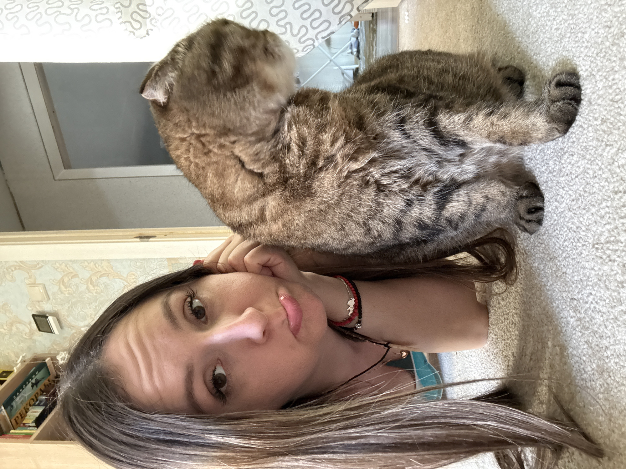 A young woman with long, brown hair is lying on a carpeted floor, resting her head on her hand and making a pouty face. Beside her, a tabby cat sits with its back to the camera, facing away from the woman. The background shows a bookshelf with books and a doorway. The scene is indoors, suggesting a residential setting. The location is identified as Kroyanskoye, Russia.