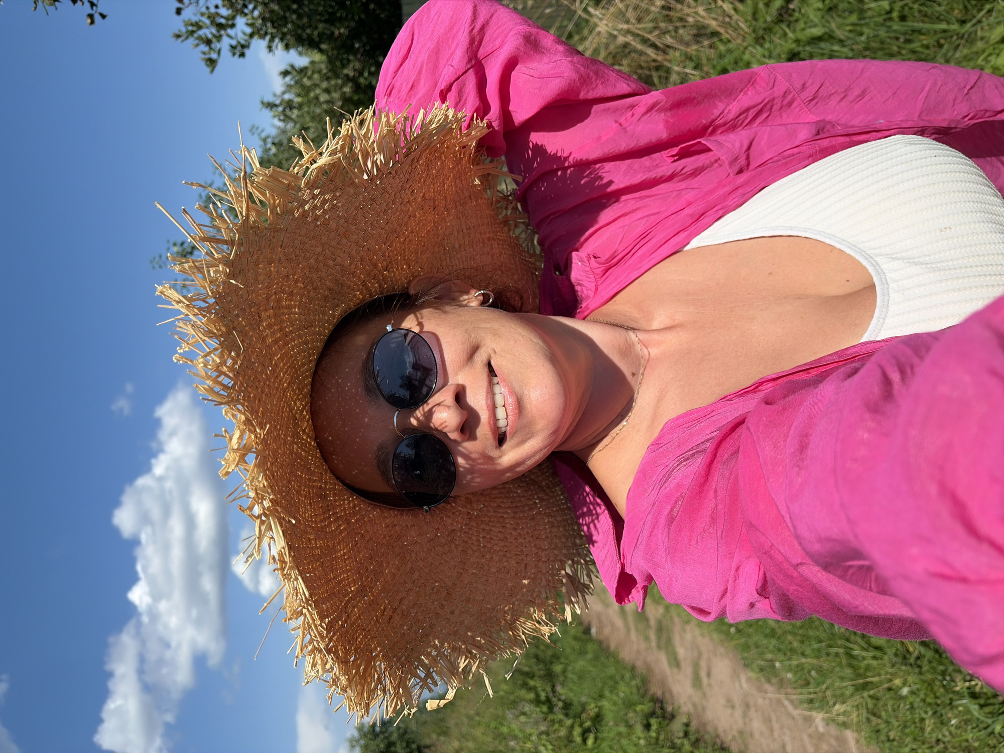 A woman is taking a selfie outdoors in a rural setting, likely in or near the village of Olkhovka, Russia, given the context. She is wearing a wide-brimmed straw hat, sunglasses, and a bright pink open shirt over a white top. Her left arm is raised, holding the camera. The background shows a dirt path, green grass, and trees under a bright blue sky with scattered white clouds.