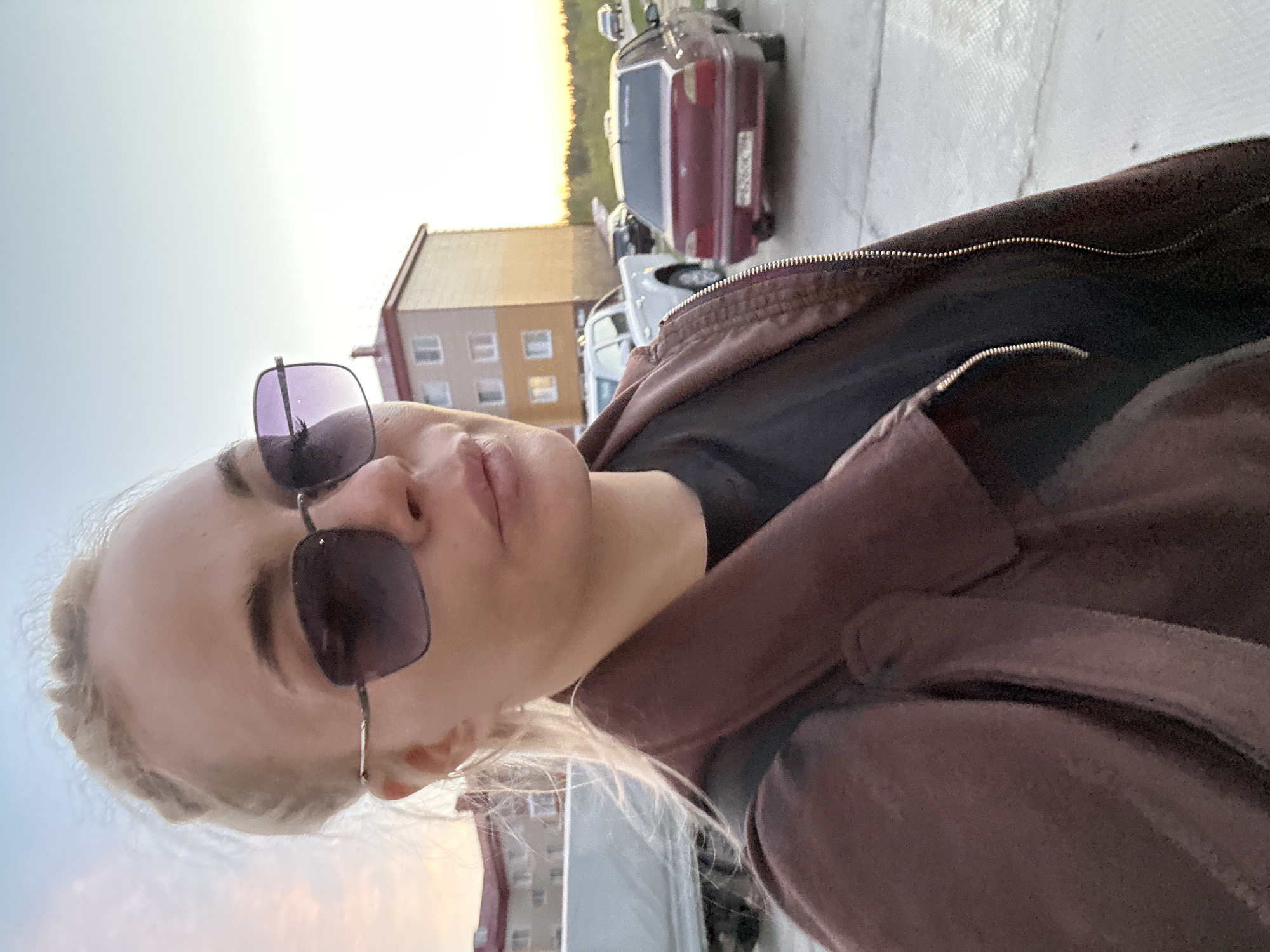 A woman wearing sunglasses and a brown jacket poses for a selfie. In the background are apartment buildings, cars, and a street. The location is Высокий, Russia.