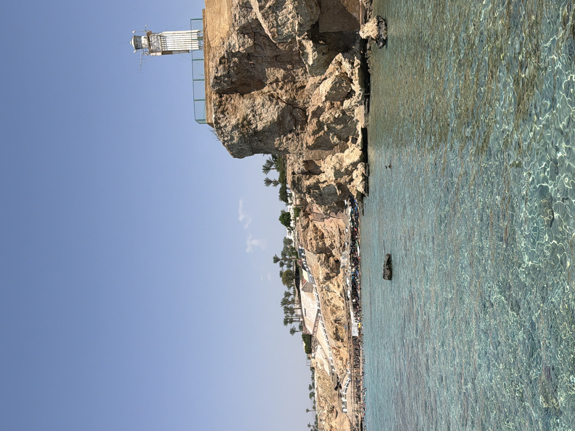 This is a high-angle, wide shot of a coastal cliffside resort area in Sharm El Sheikh, Egypt, on a clear, sunny day. The foreground is dominated by the clear, turquoise water of the Red Sea, revealing submerged rocks and coral formations below the surface. The water is calm with gentle ripples.
The midground features a bustling beach area situated at the base of a rugged, rocky cliff. Numerous beach umbrellas and loungers are visible, along with many people swimming in the sea, sunbathing, and walking along the shoreline. Stairs and pathways are carved into the cliff face, leading up to different levels of the resort. Palm trees and other vegetation dot the upper parts of the cliff, interspersed with white resort buildings.
Dominating the right side of the image and perched on the highest point of the cliff is a white lighthouse. It is a tall, slender structure with a lantern room at the top and various antennas attached. A green railing encloses the area around the lighthouse. The cliff itself is characterized by its stratified rock formations, appearing dry and arid, typical of the region. The sky is a clear, bright blue with no clouds.