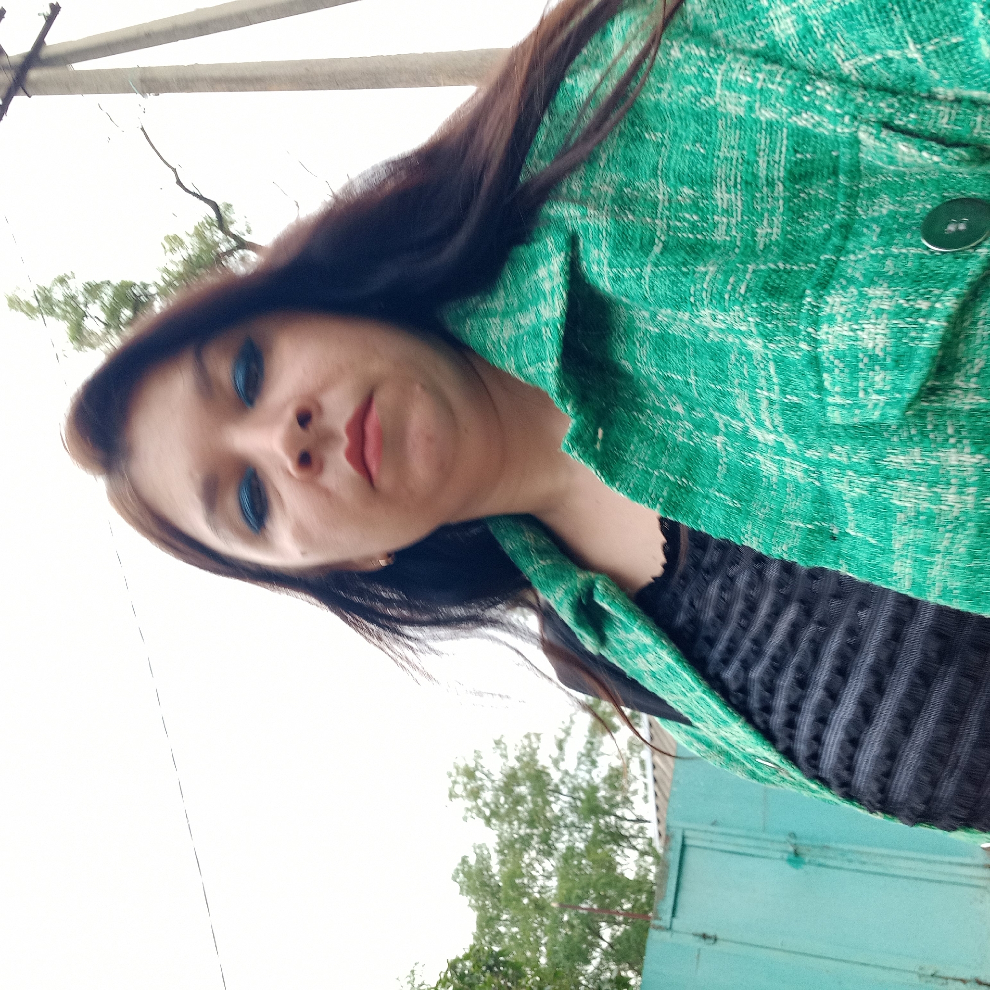 A woman is captured in a close-up, low-angle selfie outdoors. She has dark brown hair and is wearing a bright green textured jacket over a black top with a textured pattern. Blue eyeshadow is visible on her eyes, and her lips are painted a reddish-pink. In the background, a light-colored, cloudy sky dominates. To the left, a teal-colored metal gate is partially visible, and behind it, green foliage from trees. To the right, a utility pole with wires stretches upwards. The overall environment suggests an urban or semi-urban setting. The image appears to have been taken in Klyntsi, Ukraine.