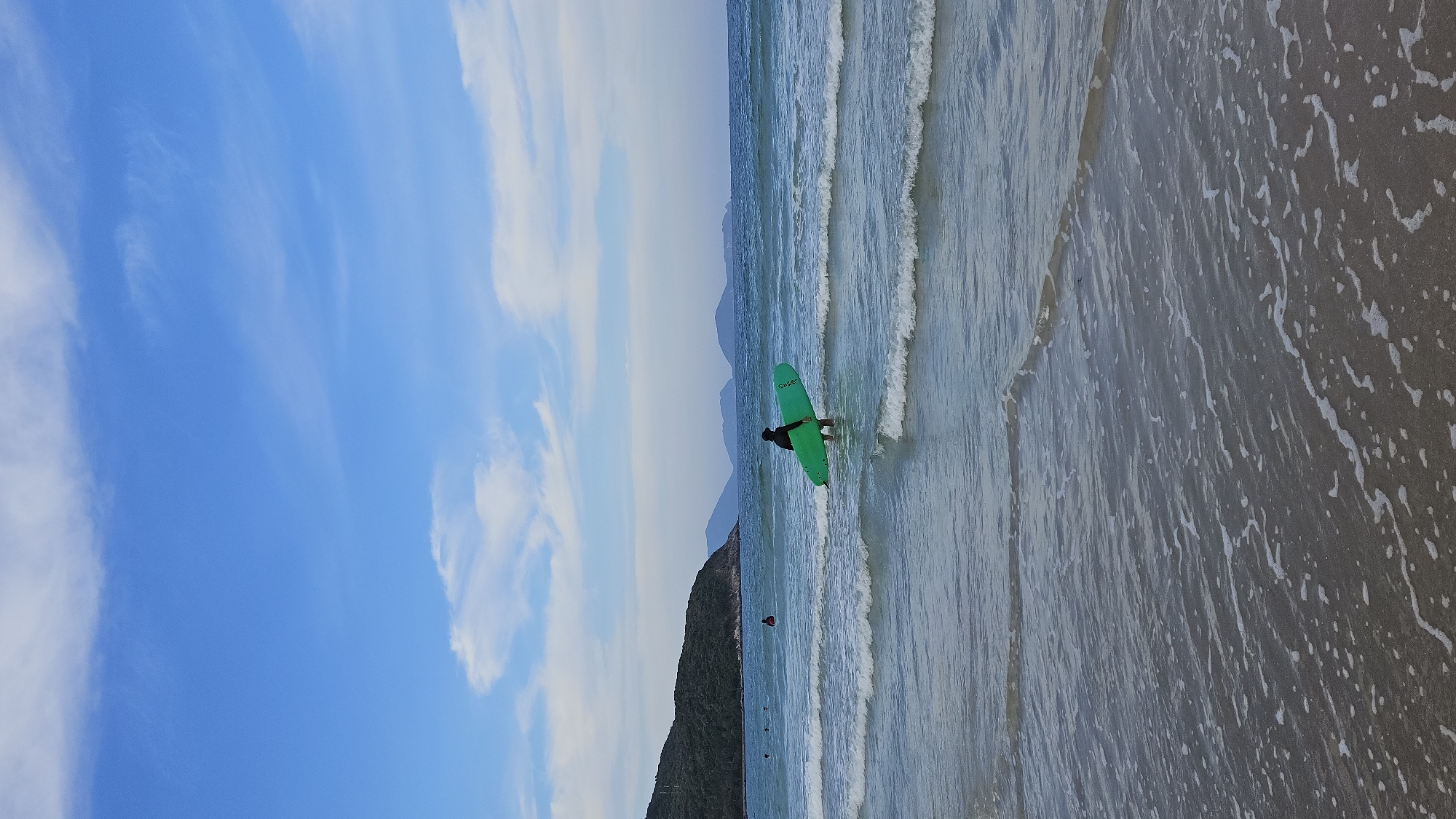 The image depicts a beach scene in Cam Lam, Vietnam. The sky is blue with scattered white clouds. A person wearing dark clothing stands in the shallow water, holding a green surfboard. The sea has gentle waves. The beach consists of wet sand. In the background, there are forested hills and mountains.