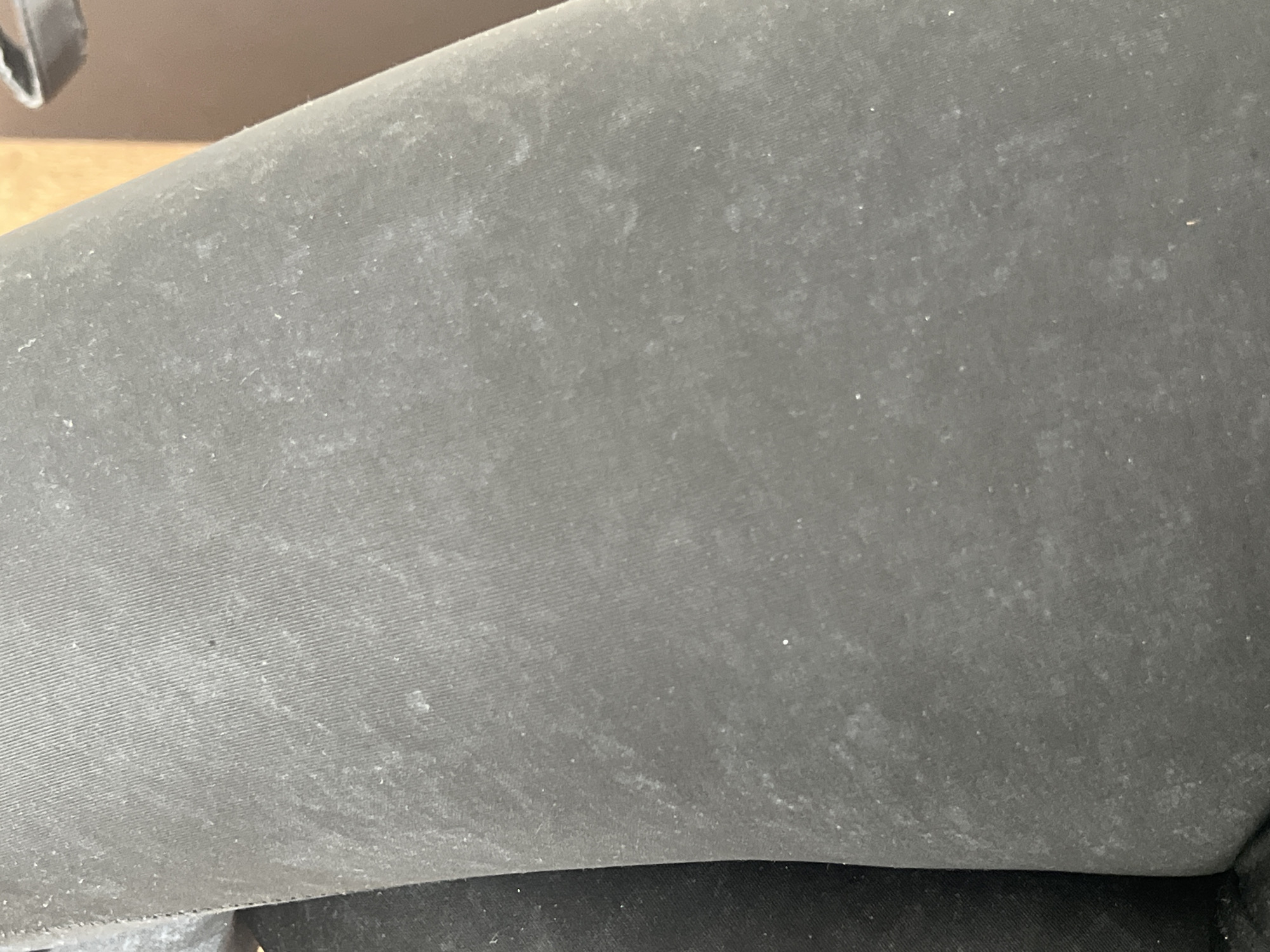 The image shows a close-up view of a dark fabric, likely leggings or pants, with a visible texture of fine lines running diagonally. The fabric appears to be slightly faded or marked with lighter-colored specks, possibly dust or lint. The background is out of focus, with a hint of a wooden surface and a dark object, possibly a bag strap, in the upper right corner. The scene is devoid of any environmental context that could identify its location in Chelyabinsk, Russia, or any other specific place. No people, actions, or interactions are depicted.