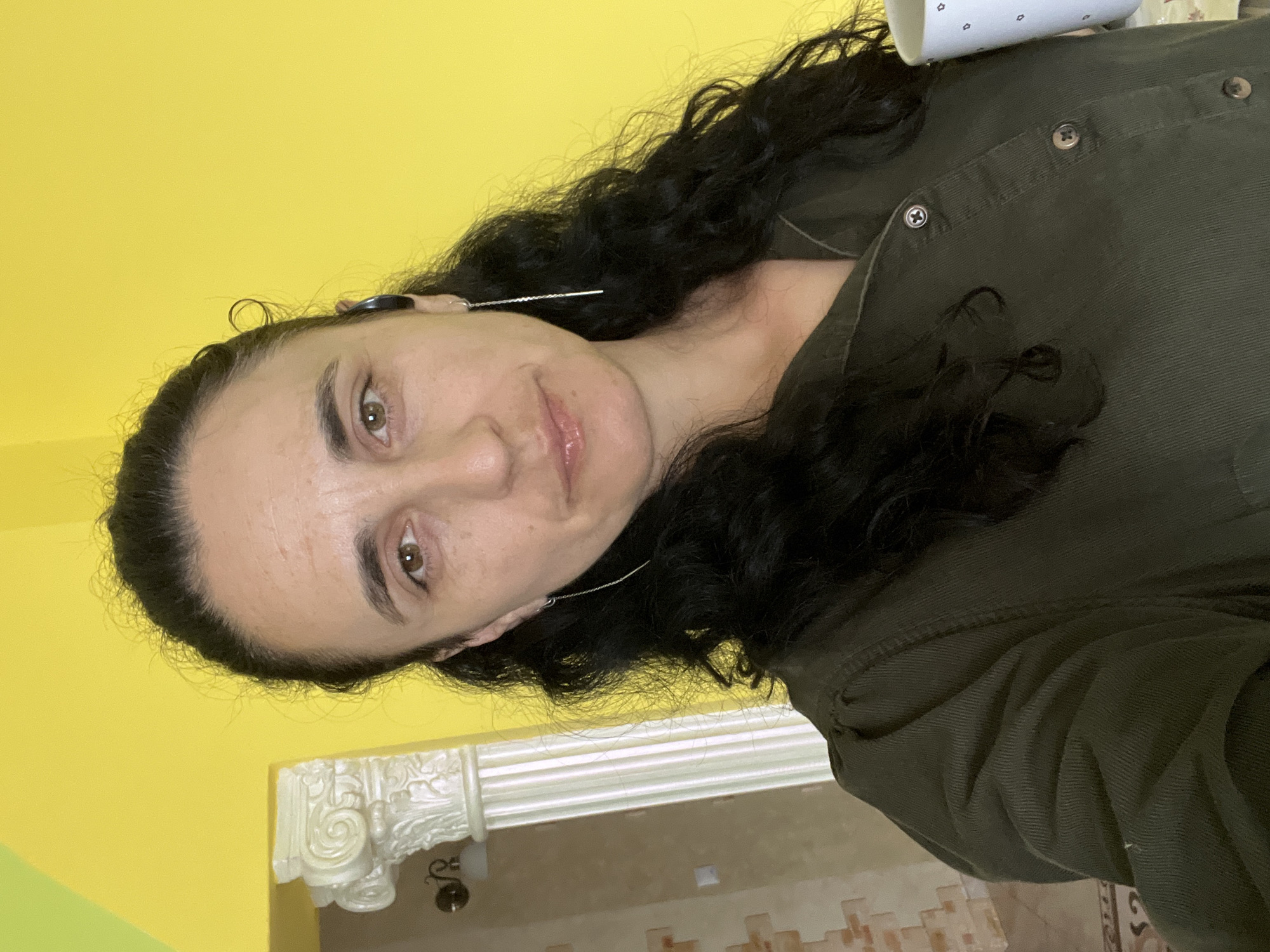 A medium shot taken from chest-level shows a woman from the chest up, holding a white mug with a blue star pattern in her right hand. She has long, dark, curly hair pulled back into a ponytail, with some strands loose around her face. She is wearing a dark olive-green collared shirt with visible buttons. She has on a single long, thin silver earring in her left ear and a black earbud in her right ear. She is looking directly at the camera with a neutral expression. The background is a bright yellow wall. To the left of the woman, a white decorative pillar with a Corinthian capital is visible, along with a wall with textured beige and brown tiles. The overall scene appears to be an indoor setting, likely a home, in the city of Onokivtsi, Ukraine.