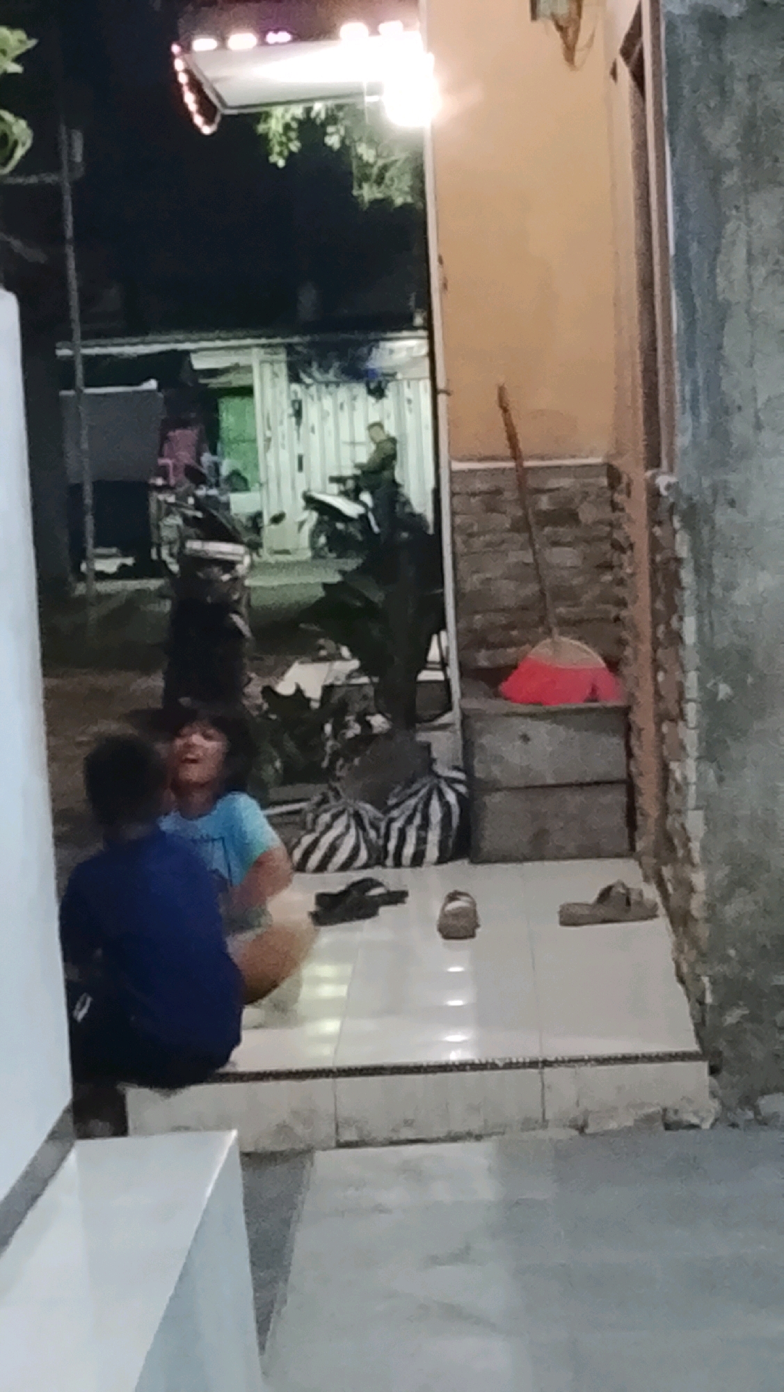 The image depicts an outdoor scene at night, likely in Ujungrusi, Indonesia. Two young individuals are seated on a tiled step in the foreground. The individual on the left is wearing a blue shirt and appears to be looking upwards and to the right. The individual on the right, seen from behind, is wearing a blue shirt. Scattered around them are several pairs of sandals, a black and white striped bag, and other bags. In the background, a person is visible near a motorcycle, and a building with a light fixture is present. A broom with a red brush is leaning against a stacked set of gray containers. The overall lighting suggests nighttime.