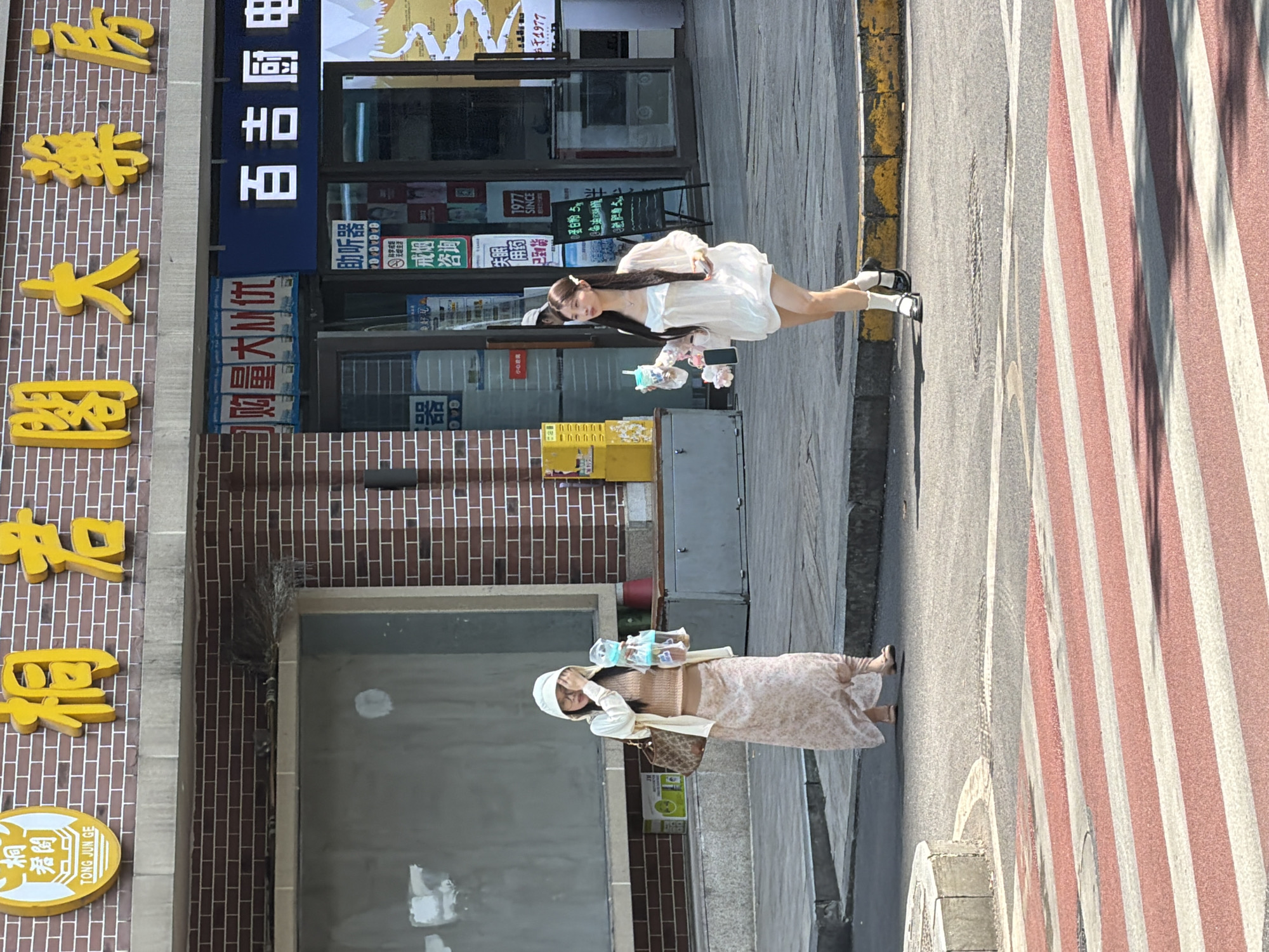 In Yuzhong District, China, two young women are walking on a sunlit street with a crosswalk in the foreground. The woman on the left is wearing a light-colored hooded jacket over a tan sweater and a long, floral print skirt. She is holding a clear plastic cup with a straw and a face mask, and her right hand is near her face. She has a patterned shoulder bag. The woman on the right is wearing a white, loose-fitting dress with a bow detail at the bust and white socks with black shoes. She is holding two clear plastic cups with straws. The background shows the brick facade of a building with Chinese signage and store entrances. A crosswalk with red and white stripes is visible in the foreground, with shadows cast across it.