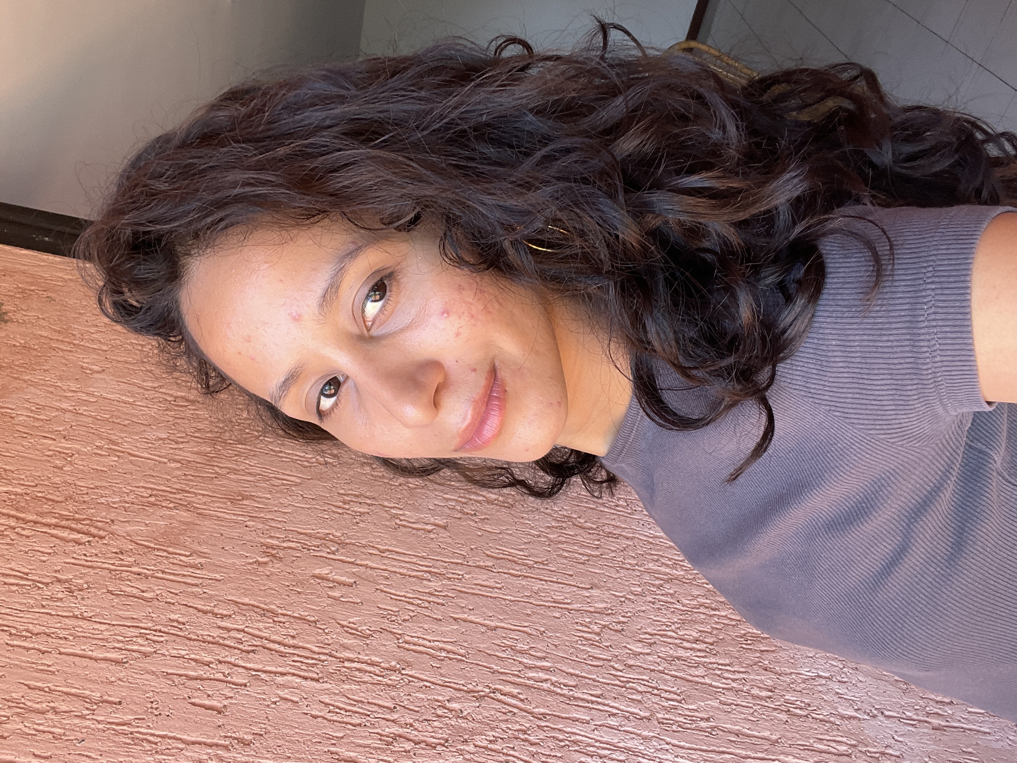 A woman with curly, dark brown hair is leaning against a textured, salmon-colored wall. She is wearing a grey, ribbed, short-sleeved top. Her face is turned towards the camera, and she has a slight smile. Her right eye is visible, as well as part of her nose and lips. There are some minor blemishes on her cheeks. A gold hoop earring is visible on her right ear. The background shows a hint of a grey floor and a doorframe in the distance, suggesting an outdoor or semi-outdoor urban setting, potentially in the city of Porlamar, Venezuela. The lighting suggests it is daytime.